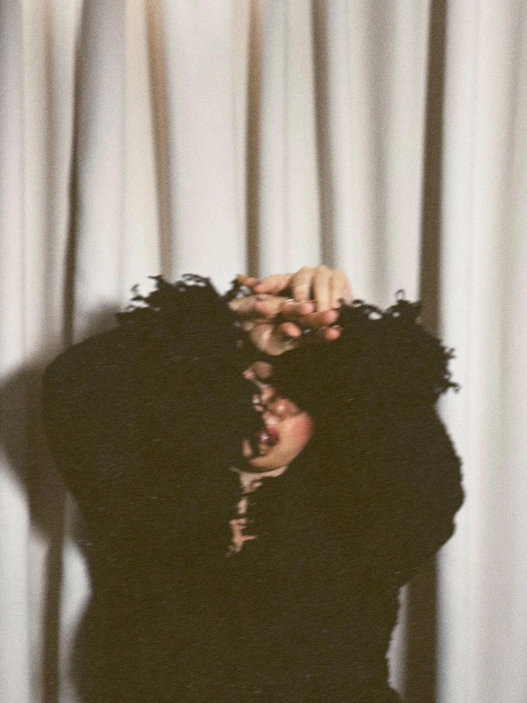 A person with curly hair, wearing makeup, holding their hand up near their face, with a neutral background.