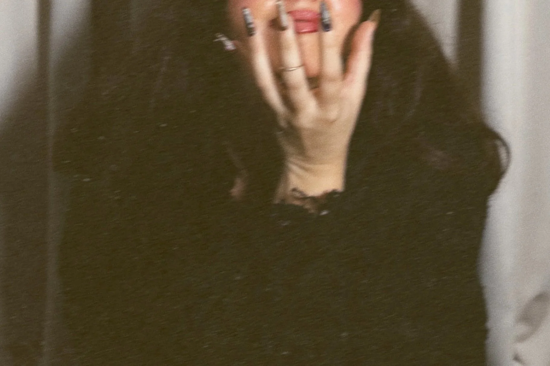 A woman taking a mirror selfie. She is wearing a black long-sleeve top and has rings on her fingers. She is covering part of her face with her hand, showing her brightly painted nails.