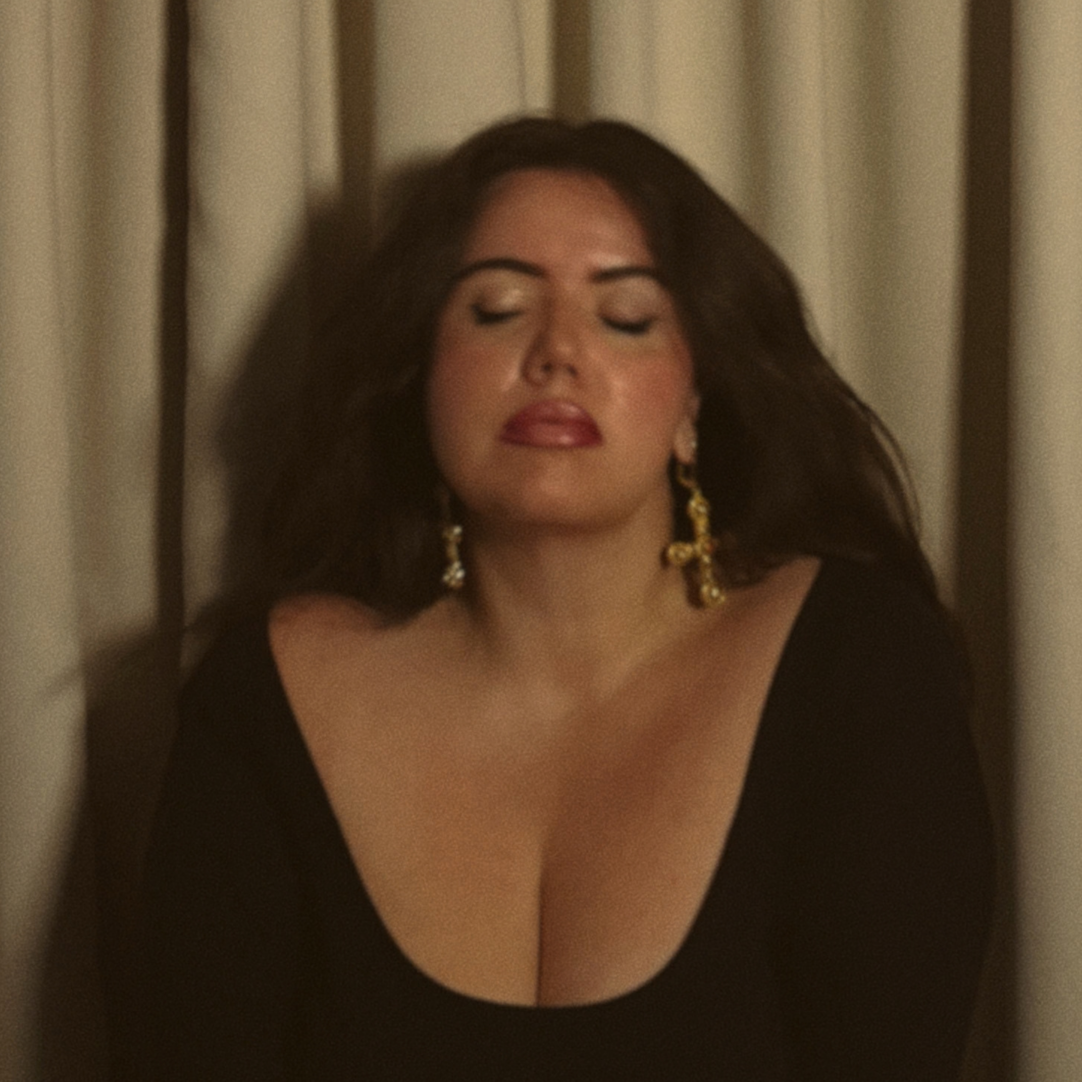 Close-up of a woman with dark hair, closed eyes, and makeup, in front of a curtain.