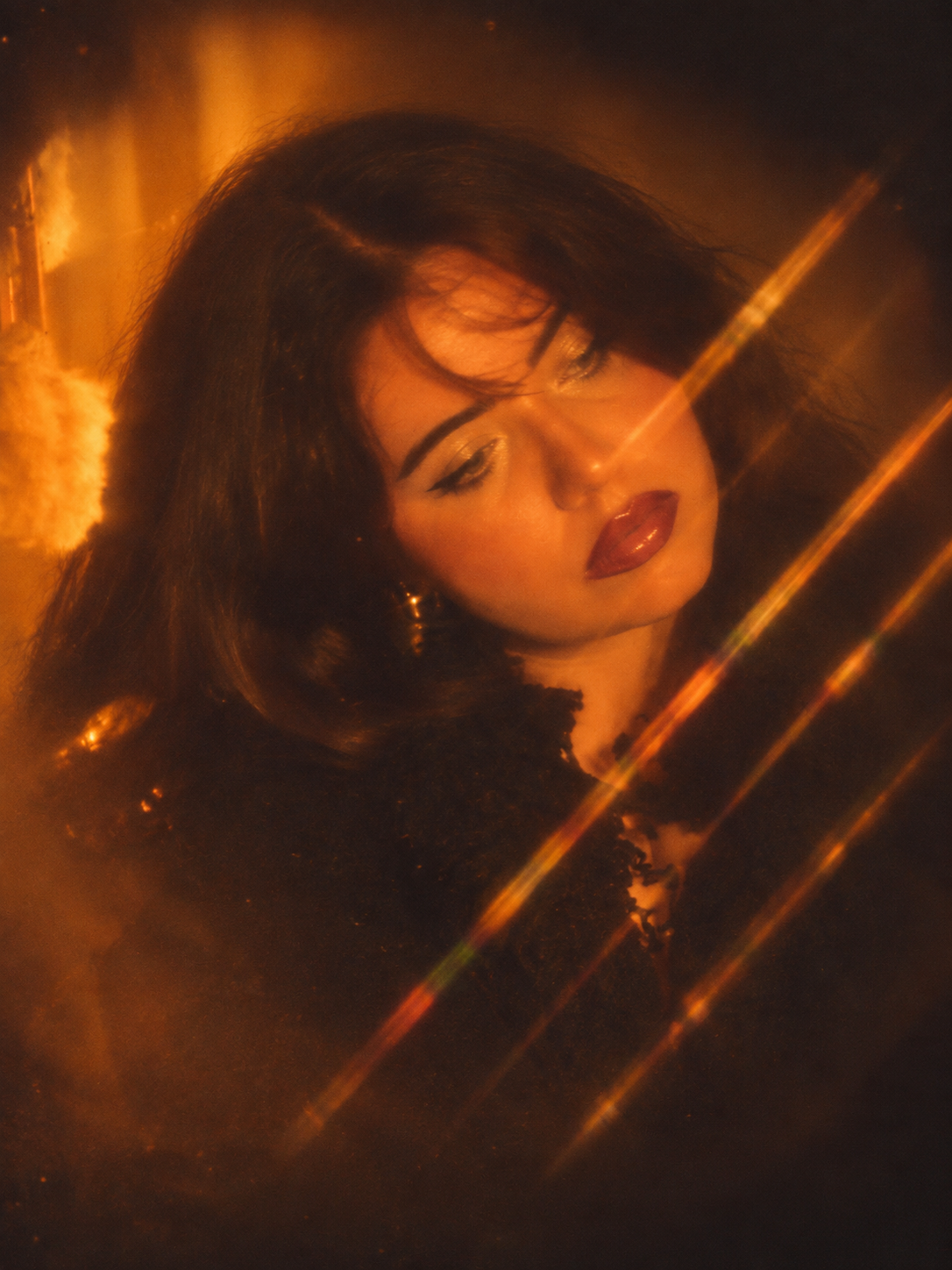 A woman with dark hair and makeup, looking downward, illuminated by warm, golden light and lens flares.