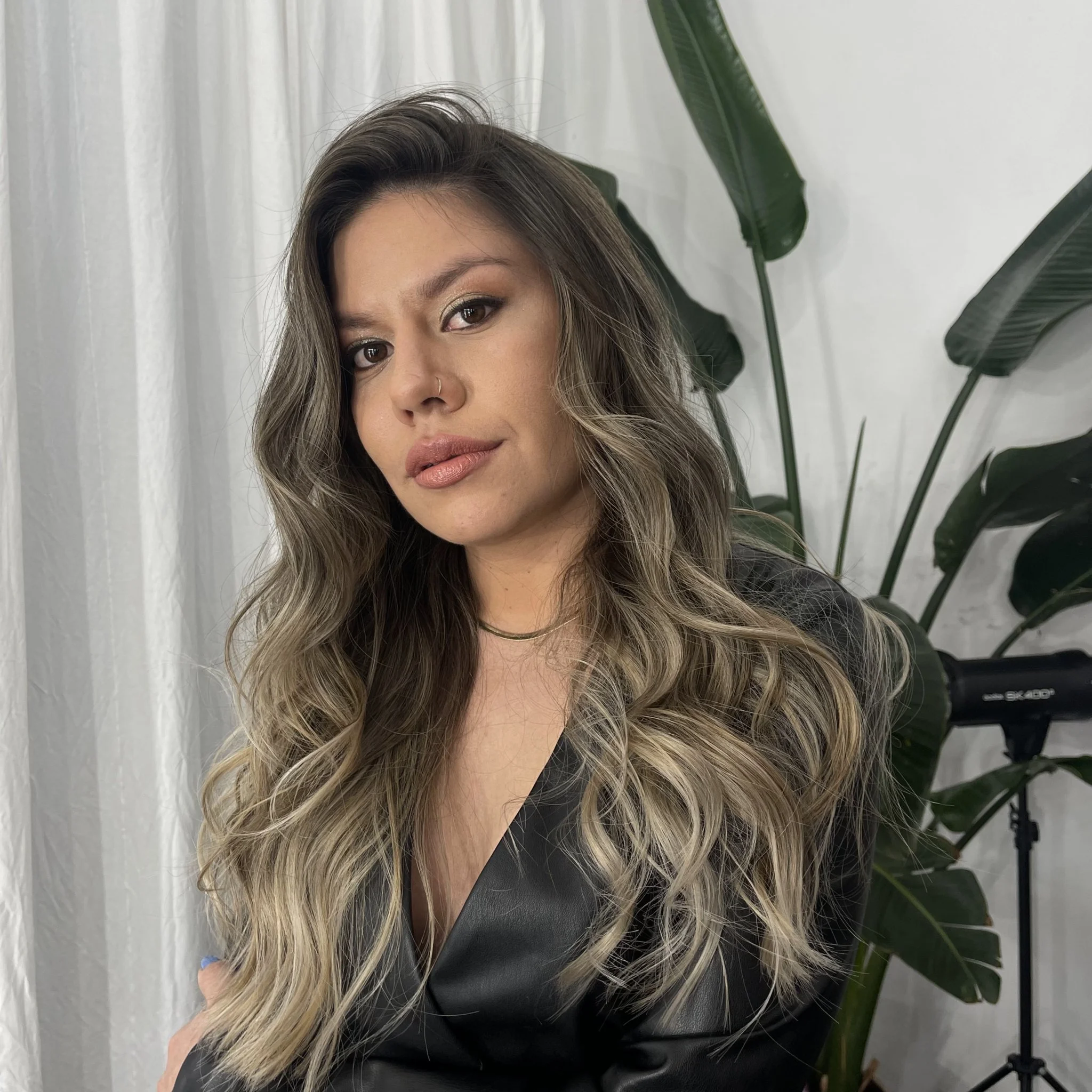 Mujer con cabello lisado y rubio con raíces oscuras, usando maquillaje y un top negro, en un entorno con plantas verdes y cortinas blancas.