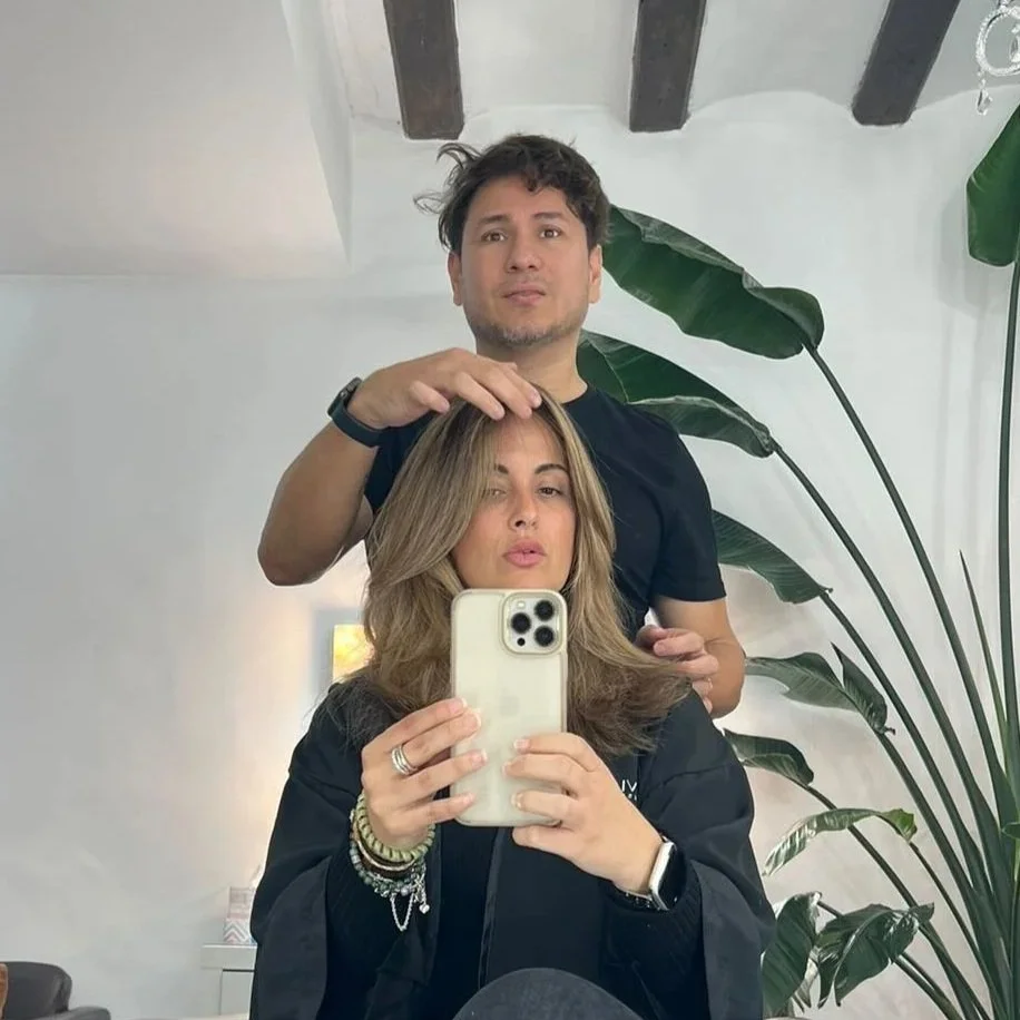 Una mujer y un hombre toman una selfie en un espejo. La mujer sostiene el teléfono y el hombre tiene la mano en la cabeza de la mujer. Detrás hay plantas grandes y una pared blanca con vigas de madera en el techo.