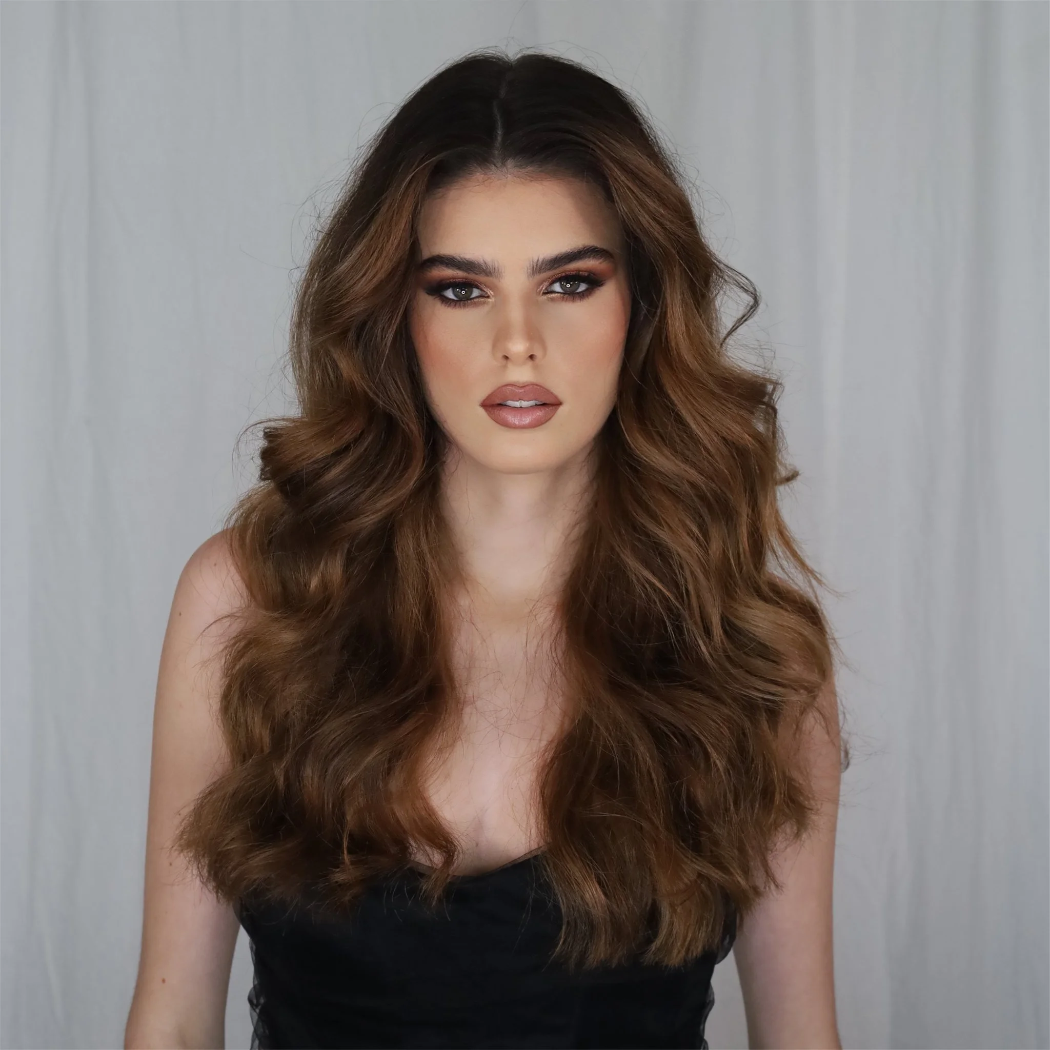 Retrato de una mujer con cabello castaño y rizado, maquillaje de ojos y labios, vestida con blusa negra, en un fondo gris claro.