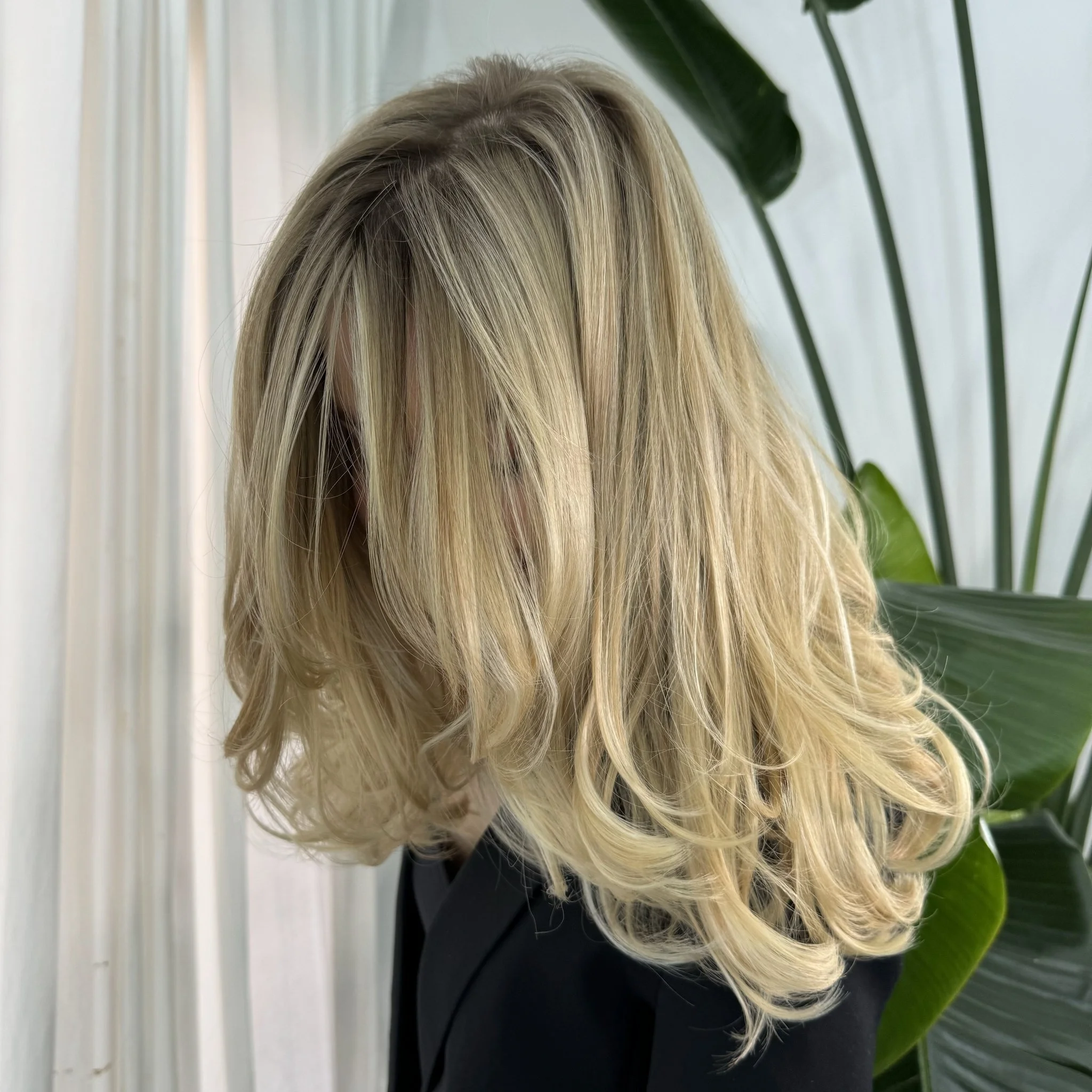Mujer con cabello rubio largo y liso, vista de perfil y de espaldas, en un entorno natural con plantas verdes.