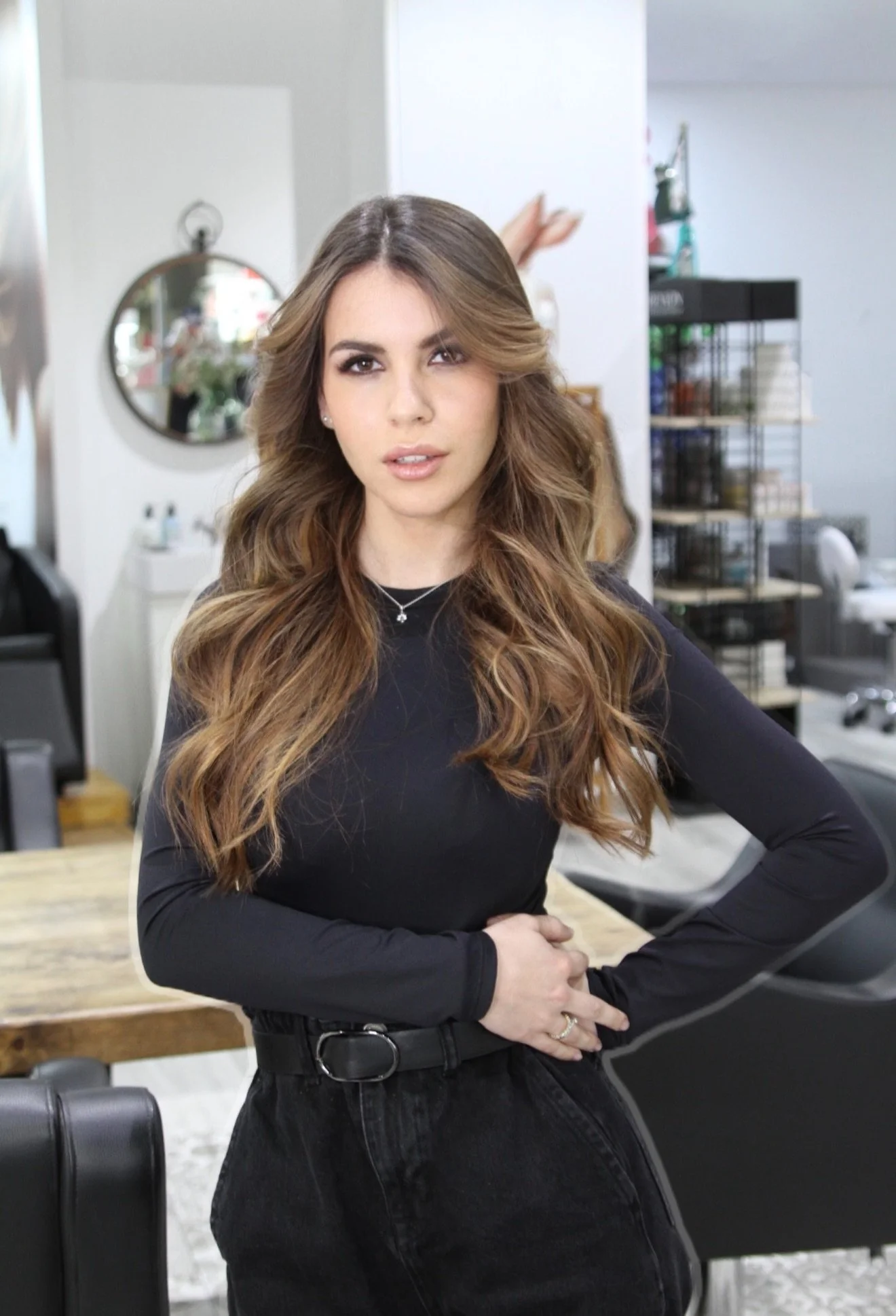 Mujer con cabello castaño largo y rizado, vestida con ropa negra, en un salón de belleza, posando con las manos en la cadera, fondo con espejo y estantes con productos