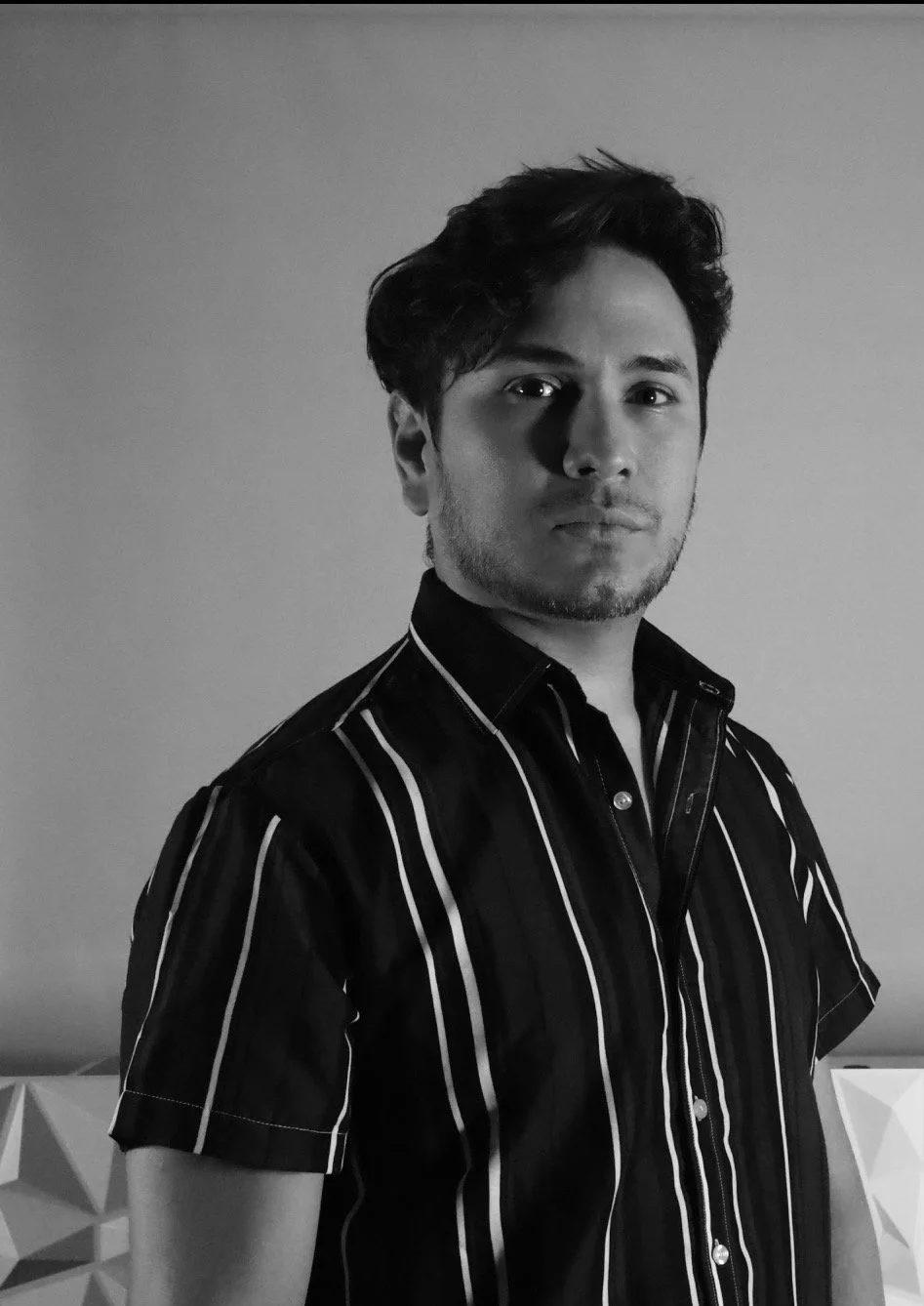Hombre joven con cabello oscuro, barba ligera, usando una camisa de rayas, de pie frente a una pared gris y con expresión seria.