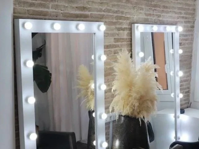 Dos espejos grandes con luces de maquillaje en un fondo de pared de ladrillos, con un florero con plumas blancas decorativas.