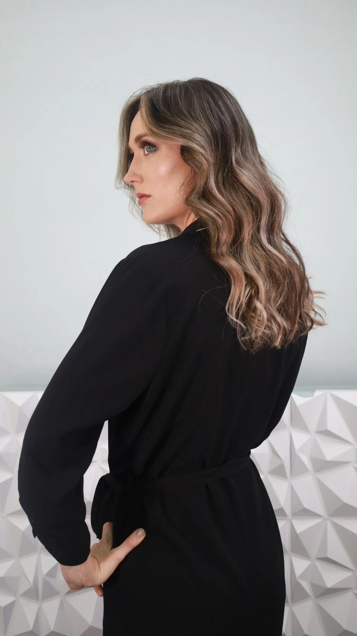 Mujer de cabello largo y rizado con maquillaje natural, usando un vestido negro con mangas largas, posando con la mano en la cadera en un fondo blanco con textura geométrica.