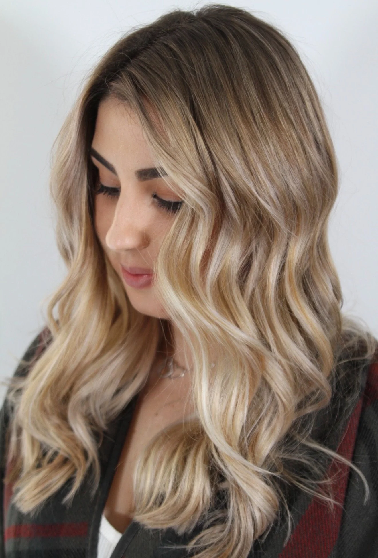 Mujer con cabello rubio con ondas, mirándose hacia abajo, con blusa blanca y chaqueta de piel en un fondo blanco.