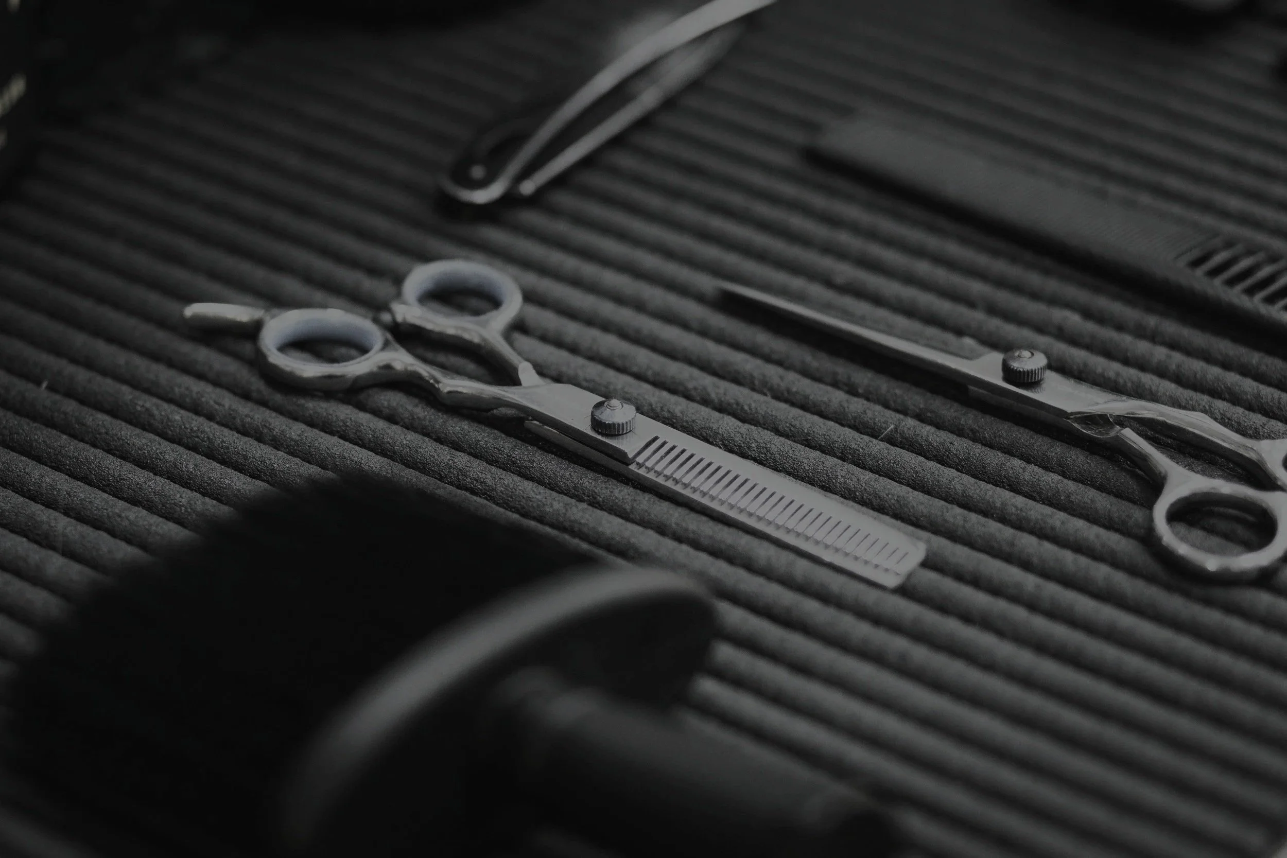 Herramientas de barbería, incluyendo tijeras, máquina de cortar cabello y navaja, colocadas sobre una superficie de goma negra.