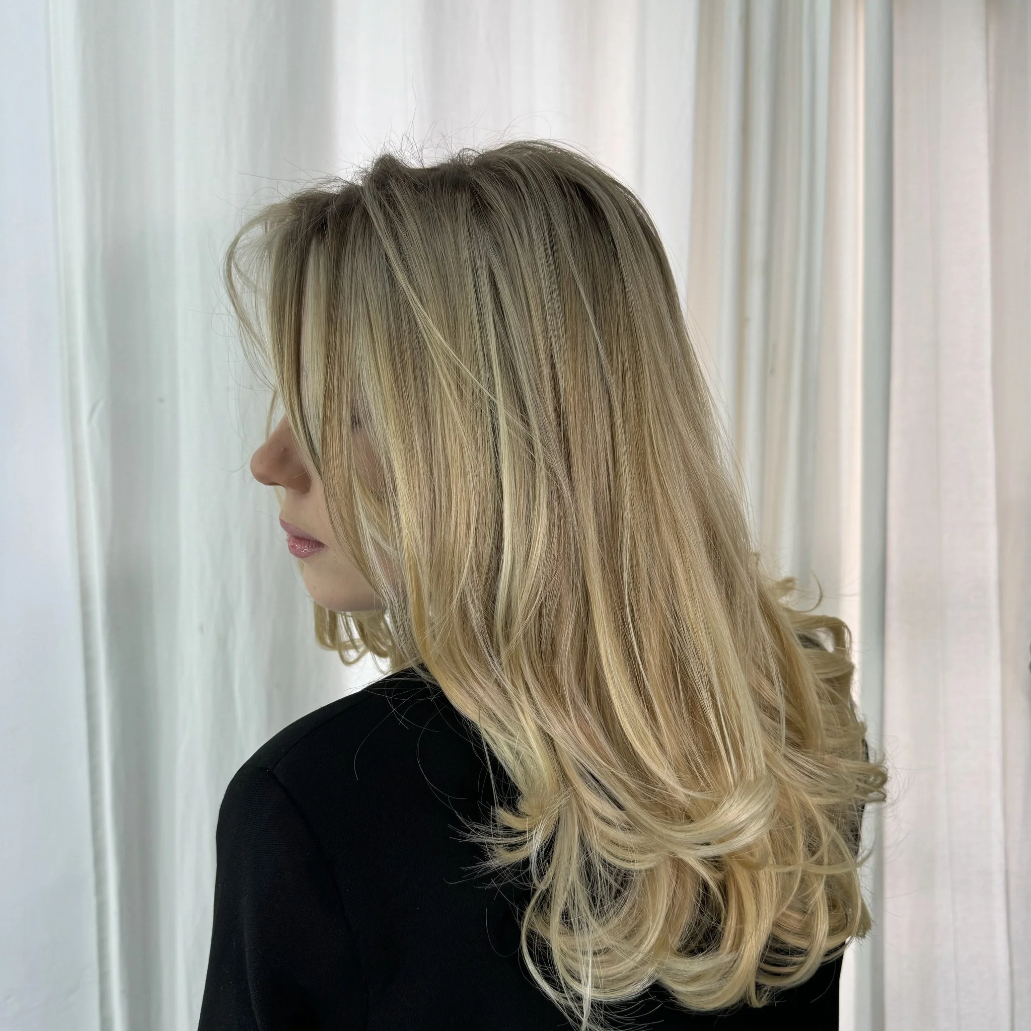 Primer plano de una mujer con cabello rubio y ondulado vista desde el lado izquierdo, con cortina beige de fondo.