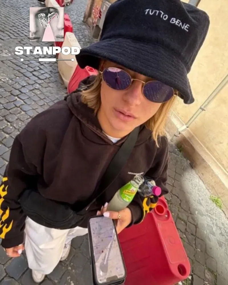 A young woman with blonde hair wearing a black bucket hat with 'TUTTO BENE' embroidered on it, purple sunglasses, and a dark hoodie with yellow accents on the sleeves. She is holding a smartphone, a can of drink, and a small bottle. She is standing on a cobblestone street near a red plastic container. In the background, there are some cardboard boxes and a poster with black and white faces and the words 'STANPOD' on the street.