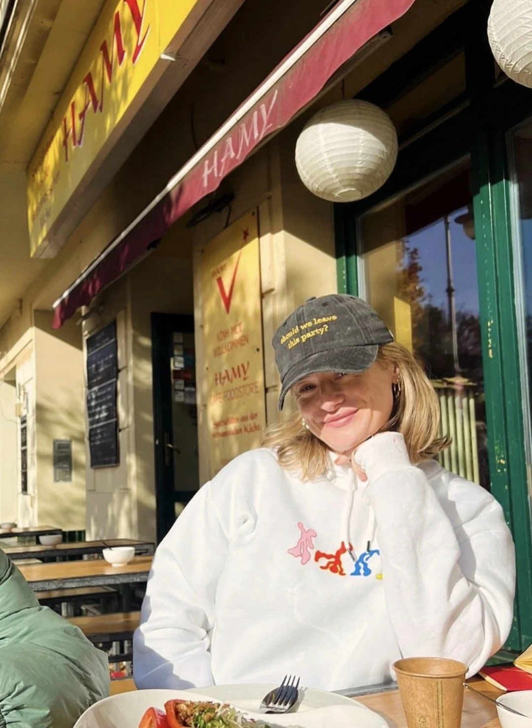 A smiling woman sits outside a restaurant, wearing a white hoodie and a dark baseball cap with yellow text, in a sunny setting with food on the table.