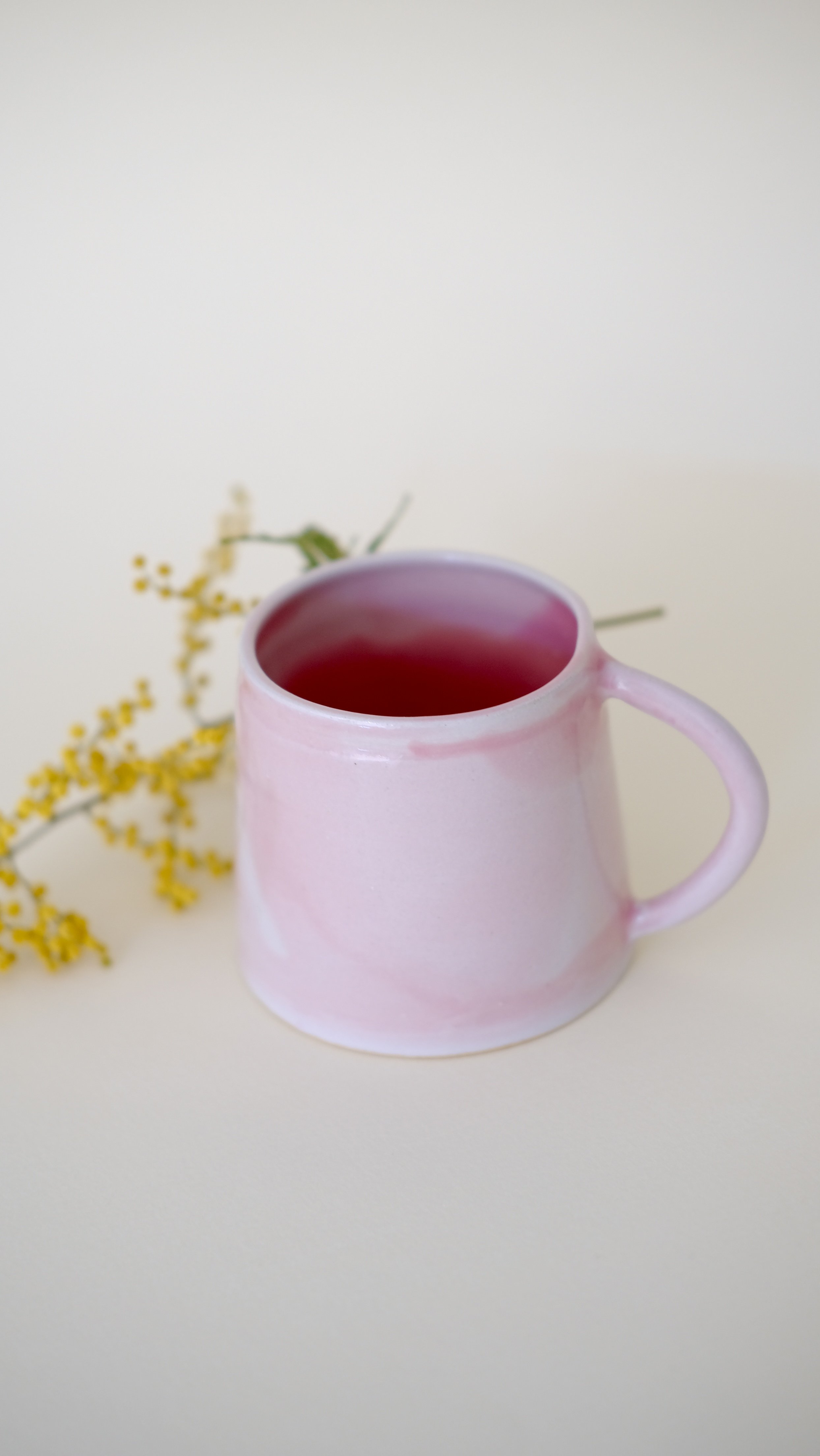 Une tasse en céramique de couleur rose pâle avec une interior violet, placée sur une surface claire avec des fleurs jaunes en arrière-plan.