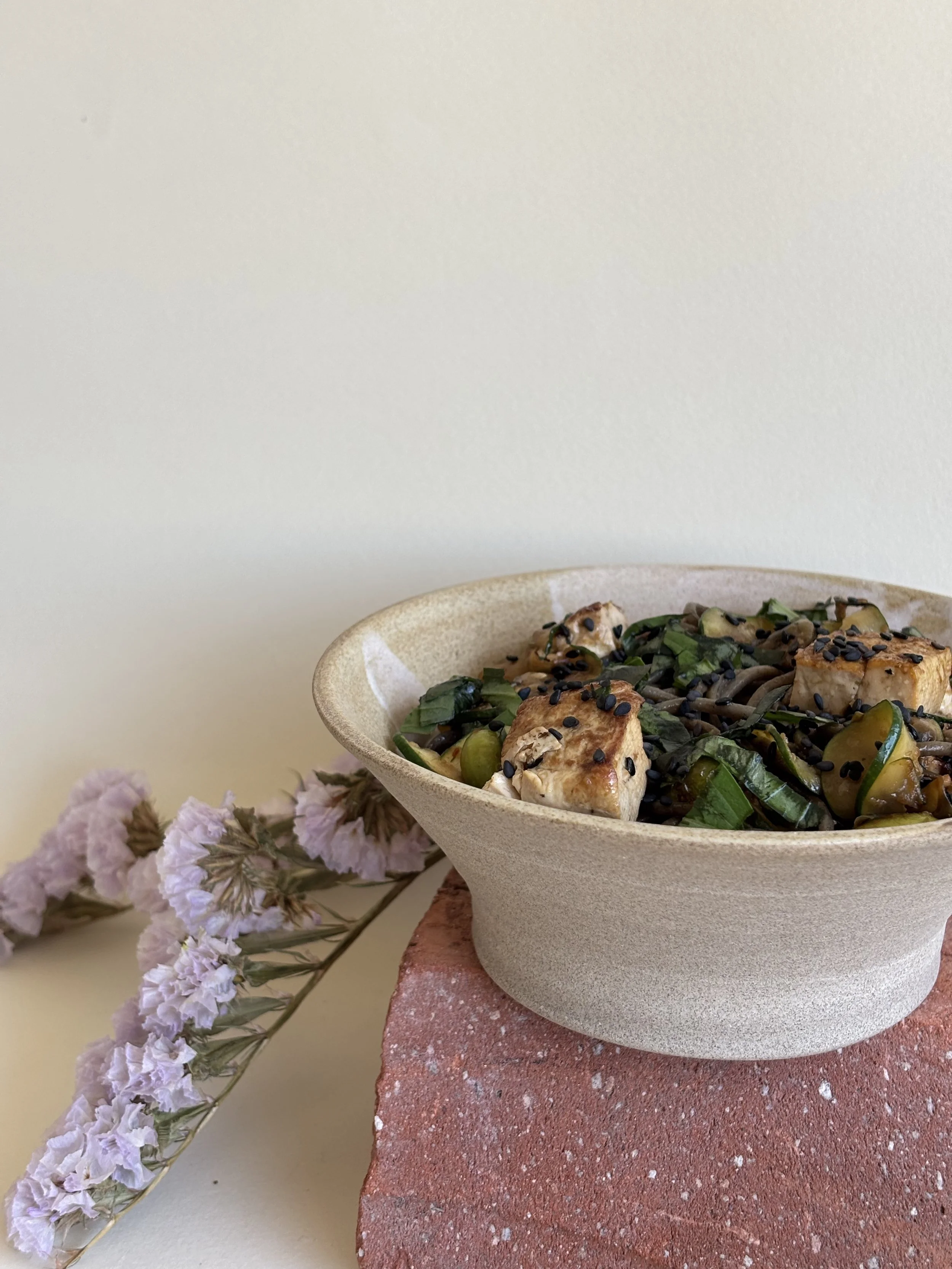 Salade dans un bol en céramique posé sur une plateforme en brique, accompagnée de fleurs violettes sèches sur un fond jaune.