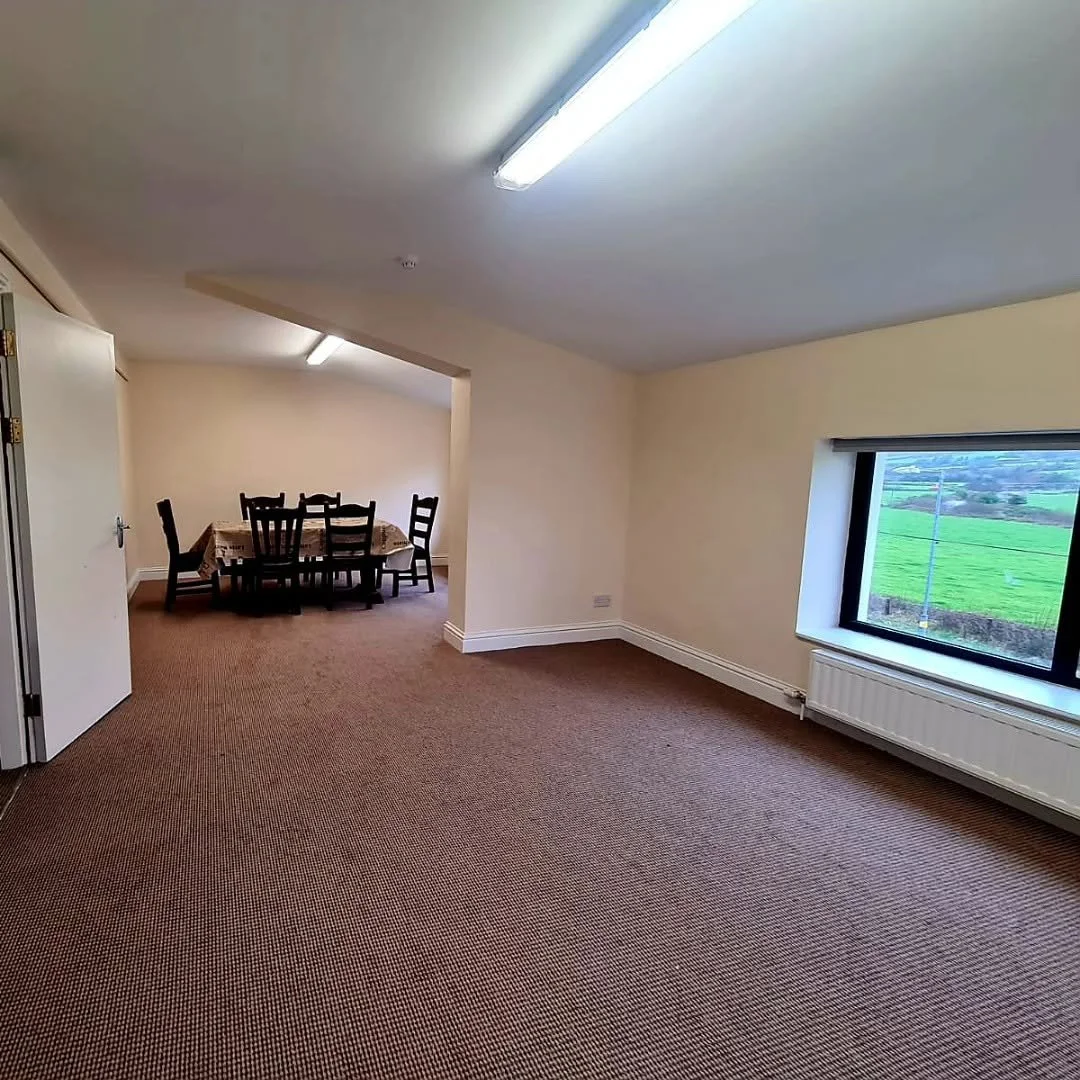 Empty room with beige walls, carpeted floor, a window showing green fields outside, and a dining area with a table and chairs.