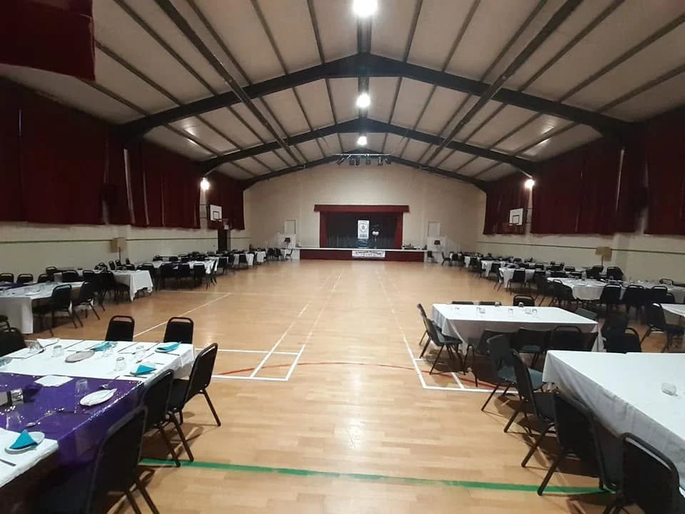 Empty multipurpose gymnasium with long tables set up for a banquet, some tables with purple tablecloths, on a hardwood floor, with a stage at the far end and basketball hoops on the sides.