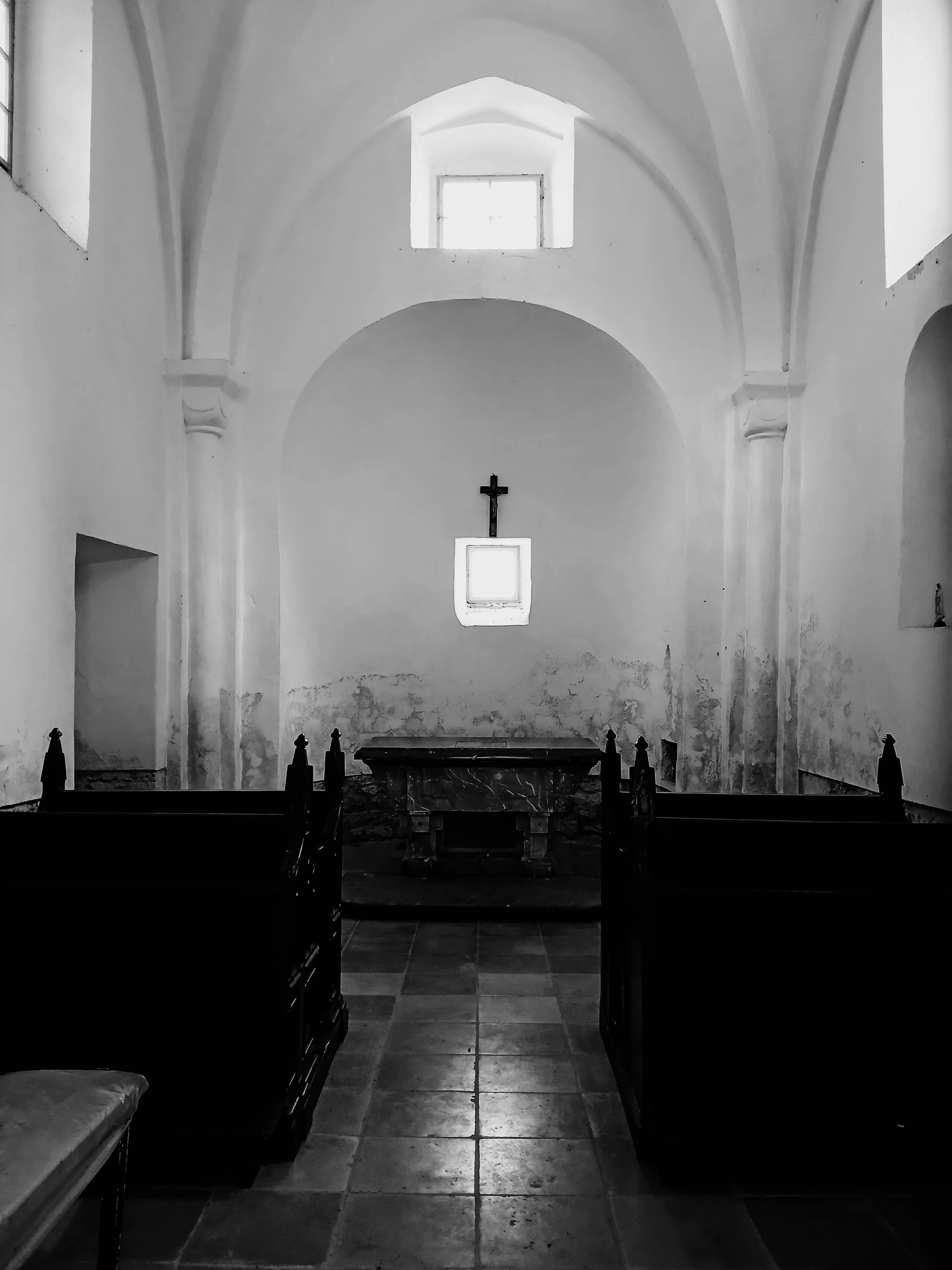 Black and white interior of a church with a high, arched ceiling, a cross on the wall at the back, and wooden pews on either side of a tiled aisle.
