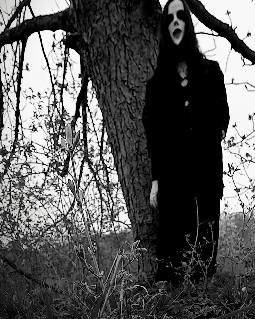 A ghost or uncanny entity with distorted facial features, standing next to a large tree in a wooded area with sparse branches and plants.