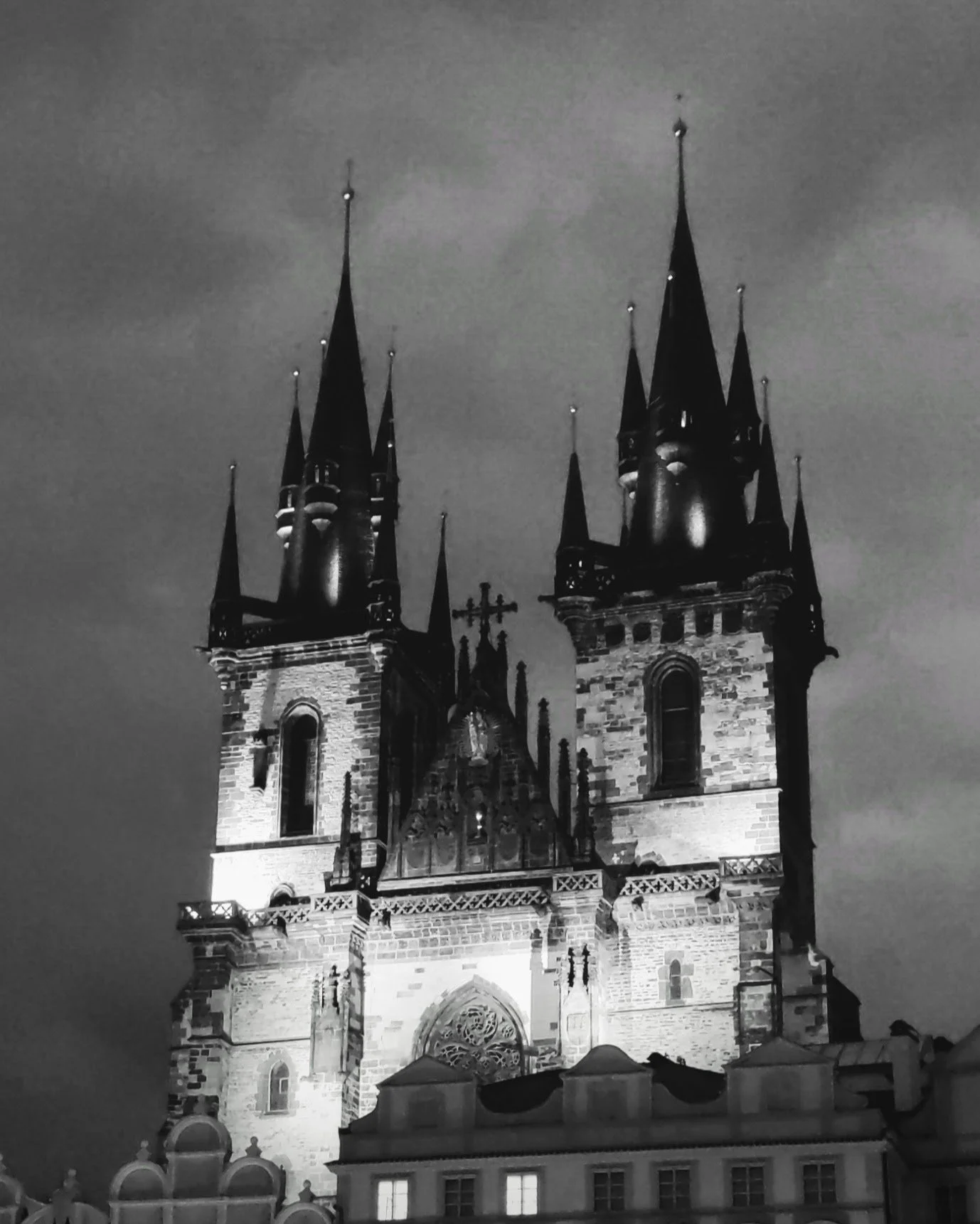 Moody nighttime view of a Gothic cathedral with tall spires and detailed stone architecture, illuminated against a dark, cloudy sky.