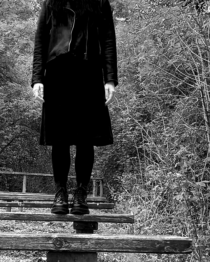 A person with abnormally long arms standing on a wooden bench in a wooded area, wearing a black leather jacket, black skirt, black tights, and black combat boots.