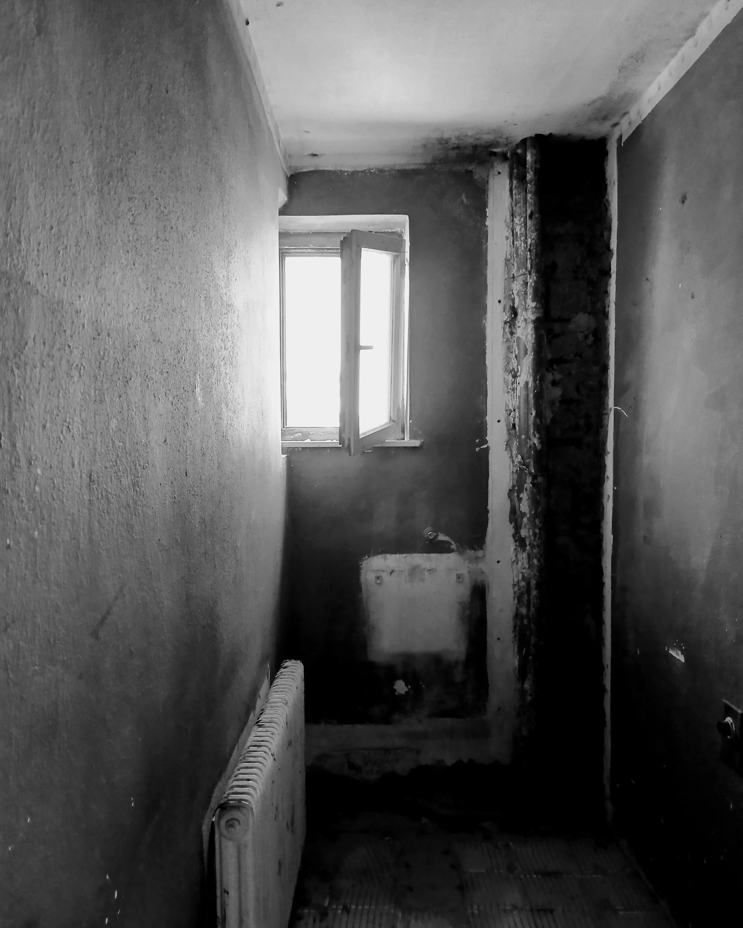An unfinished room in an abandoned house with a window emitting natural light, a partially removed wall revealing brick and plaster, and a radiator along one wall.