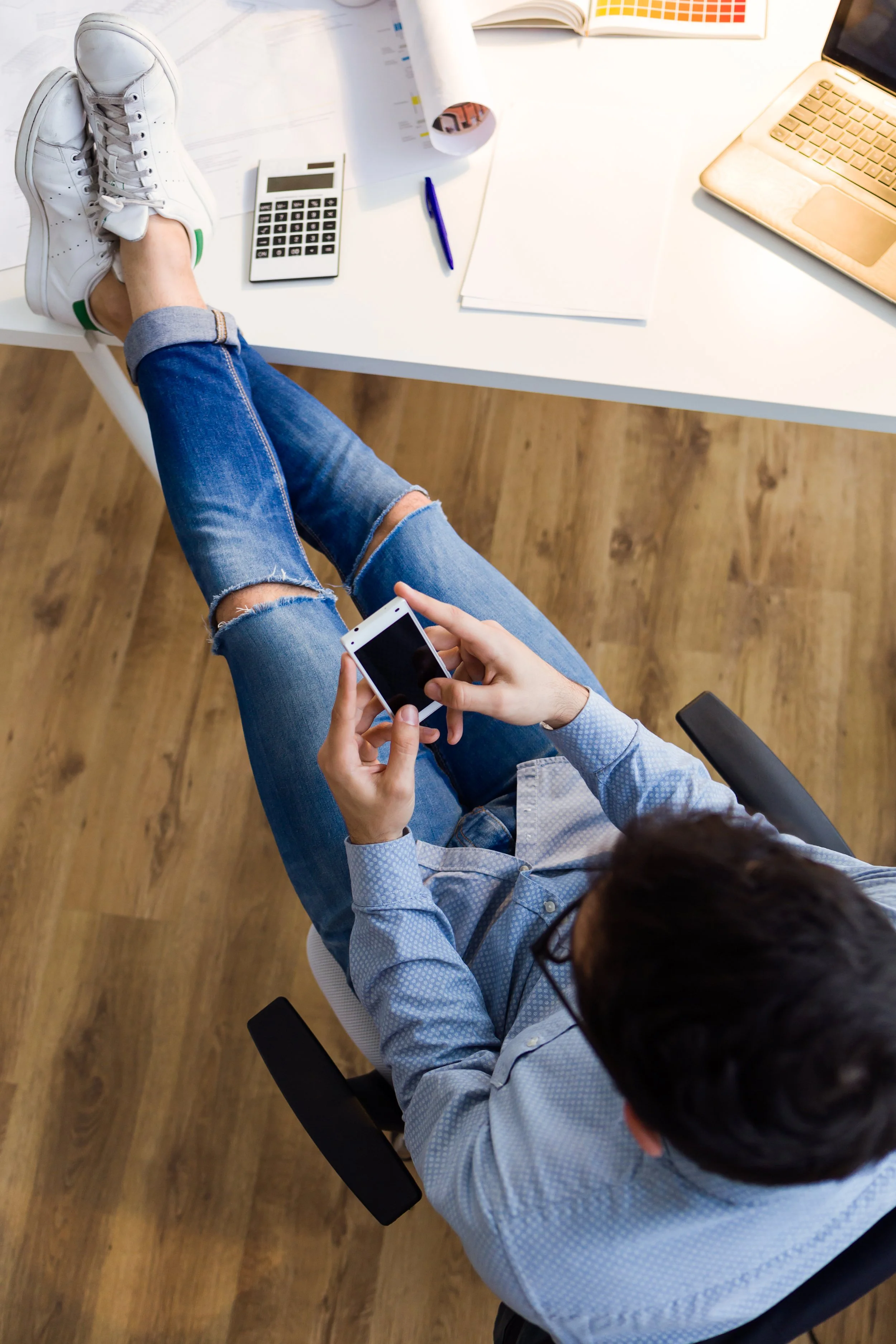 Personne assise à un bureau avec un ordinateur portable, un smartphone, une calculatrice, un stylo, des papiers et un livre ouvert sur le bureau.