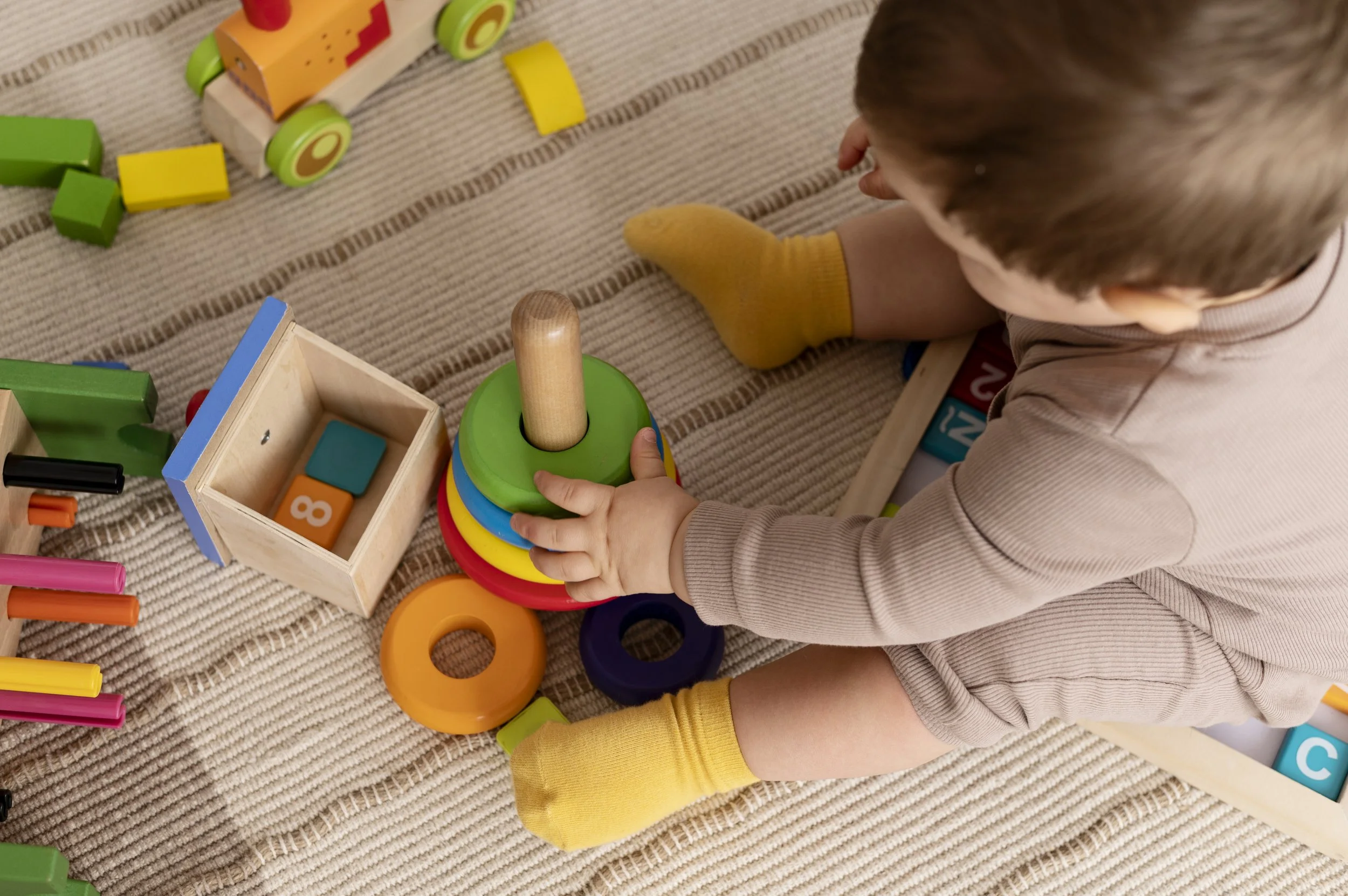 Un enfant joue avec des jouets de construction en bois, dont des formes colorées et des blocs, sur un tapis rayé beige.