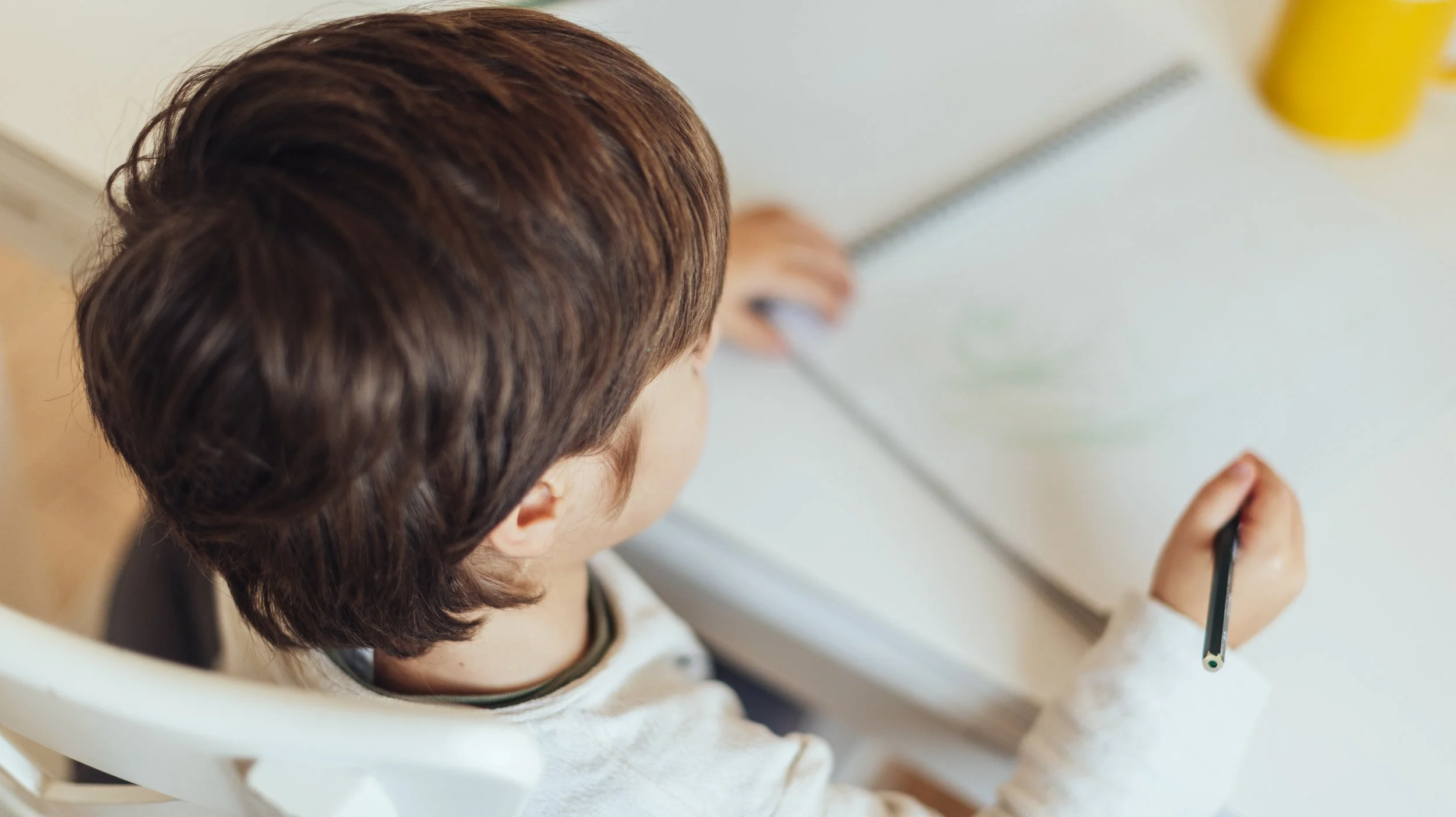 Un enfant avec des cheveux bruns courts utilise un stylo pour dessiner ou écrire sur une surface blanche, probablement un tableau ou une ardoise, dans une salle d'école ou un environnement d'apprentissage.