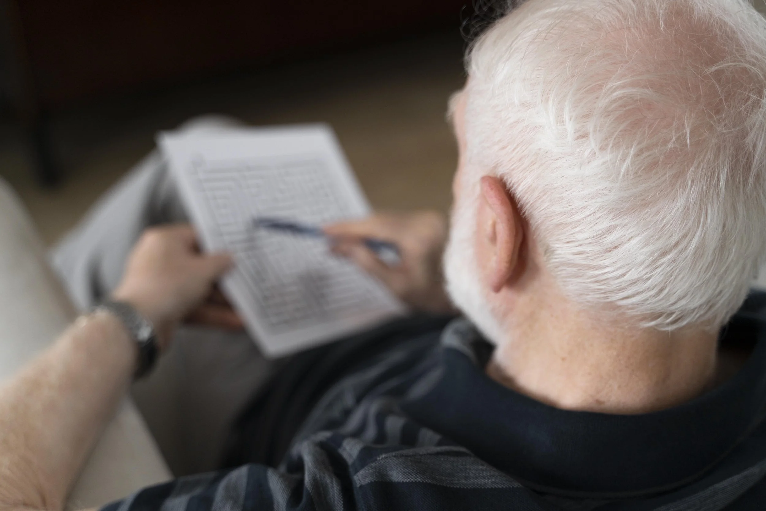 Une personne âgée avec les cheveux blancs, vue de derrière, tenant un carnet ou une fiche en main, avec un stylo, probablement en train de remplir ou de lire quelque chose.