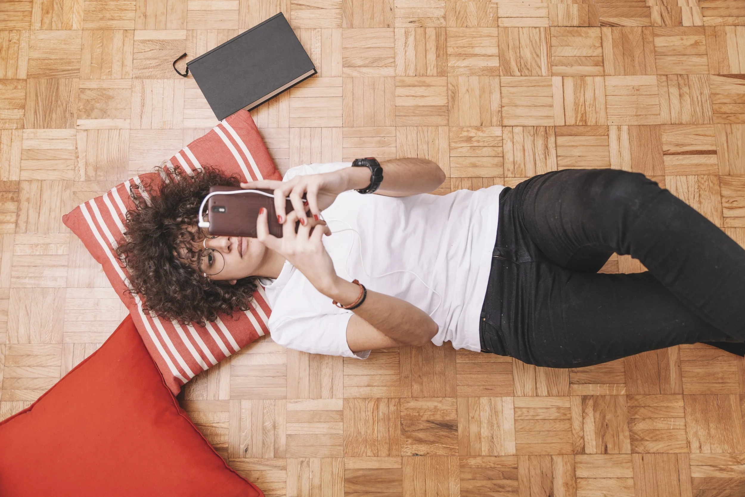 Jeune femme allongée sur un tapis avec un oreiller orange et des bandes blanches, prenant un selfie avec un téléphone. Elle porte des lunettes, un bracelet noir, un t-shirt blanc et un jean noir. Le sol est en parquet de bois.