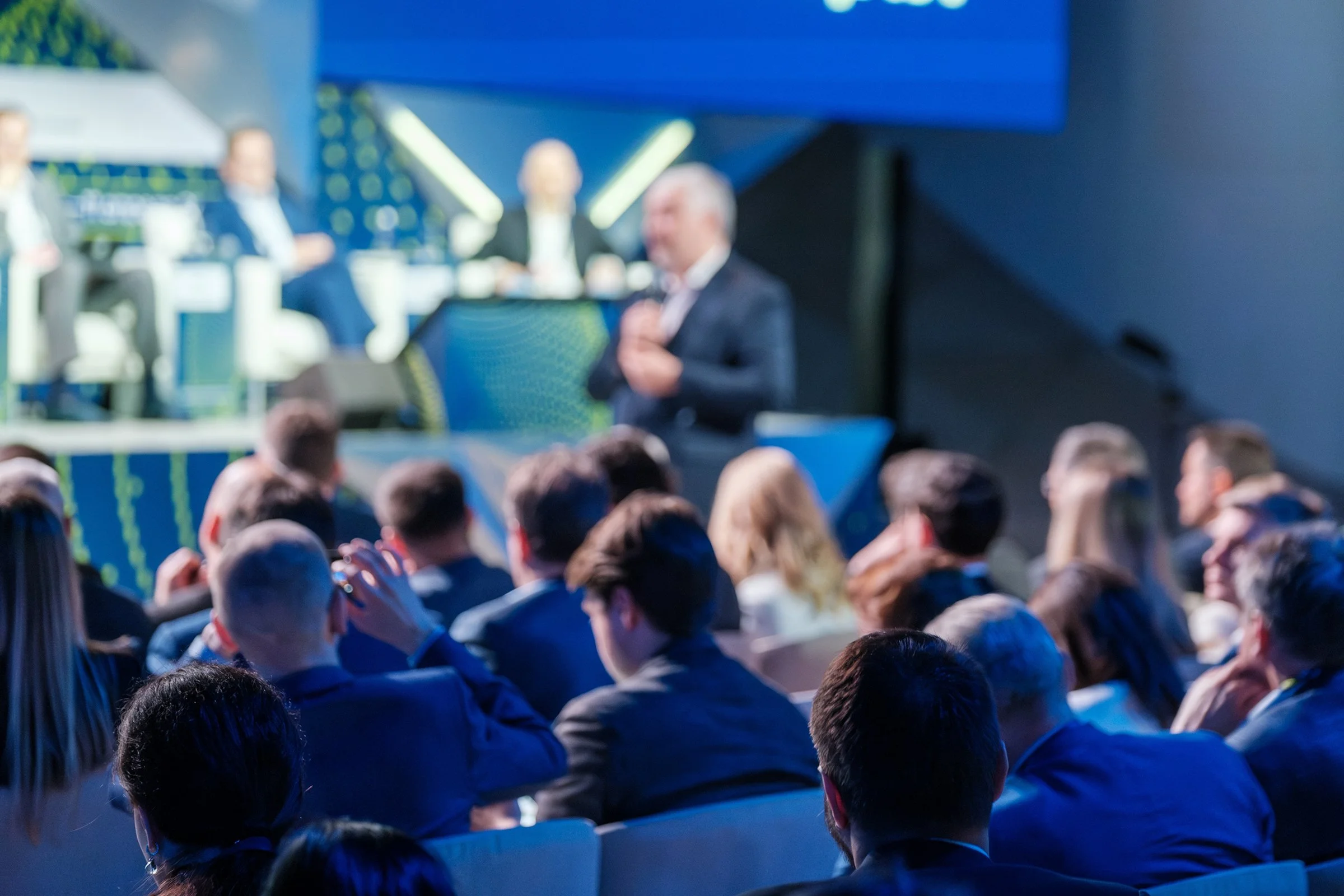 Blurred business conference with a speaker on stage and audience members seated in the foreground