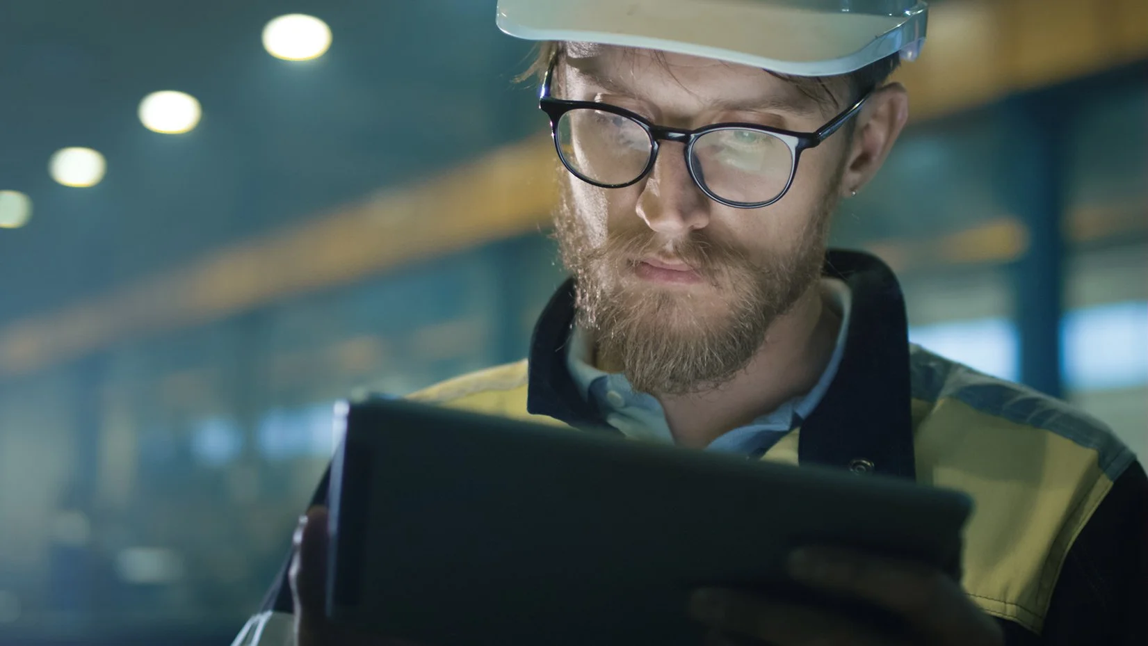 Bearded industrial engineer wearing a hard hat and safety glasses while using a tablet in a factory