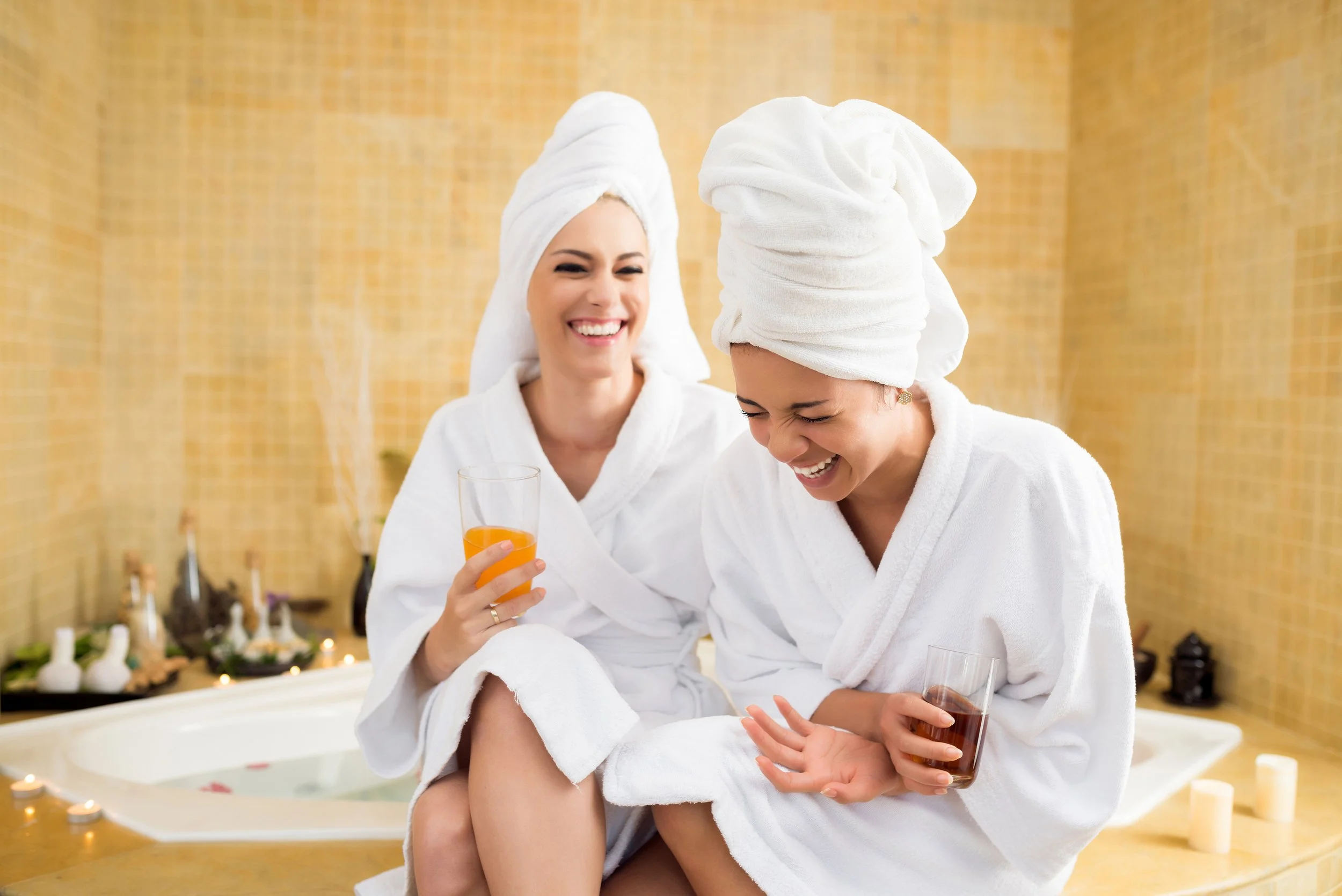 Two women in white bathrobes and towels laughing together at a spa, holding drinks beside a bathtub.