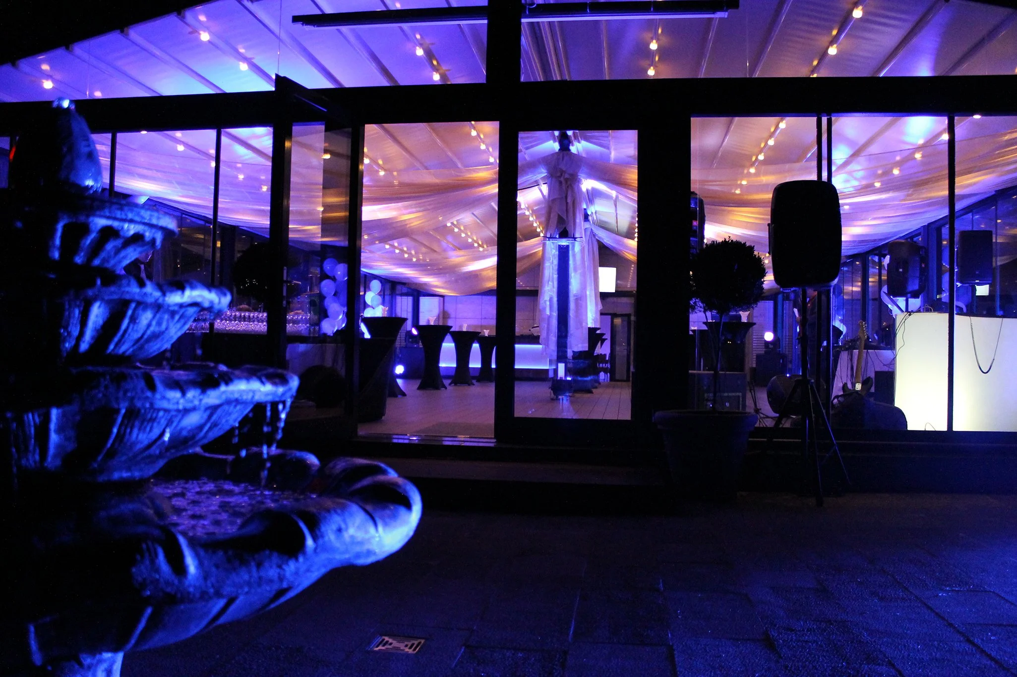 Elegant event venue illuminated in blue and purple lighting, with a tiered fountain in the foreground and a draped reception tent behind glass doors.