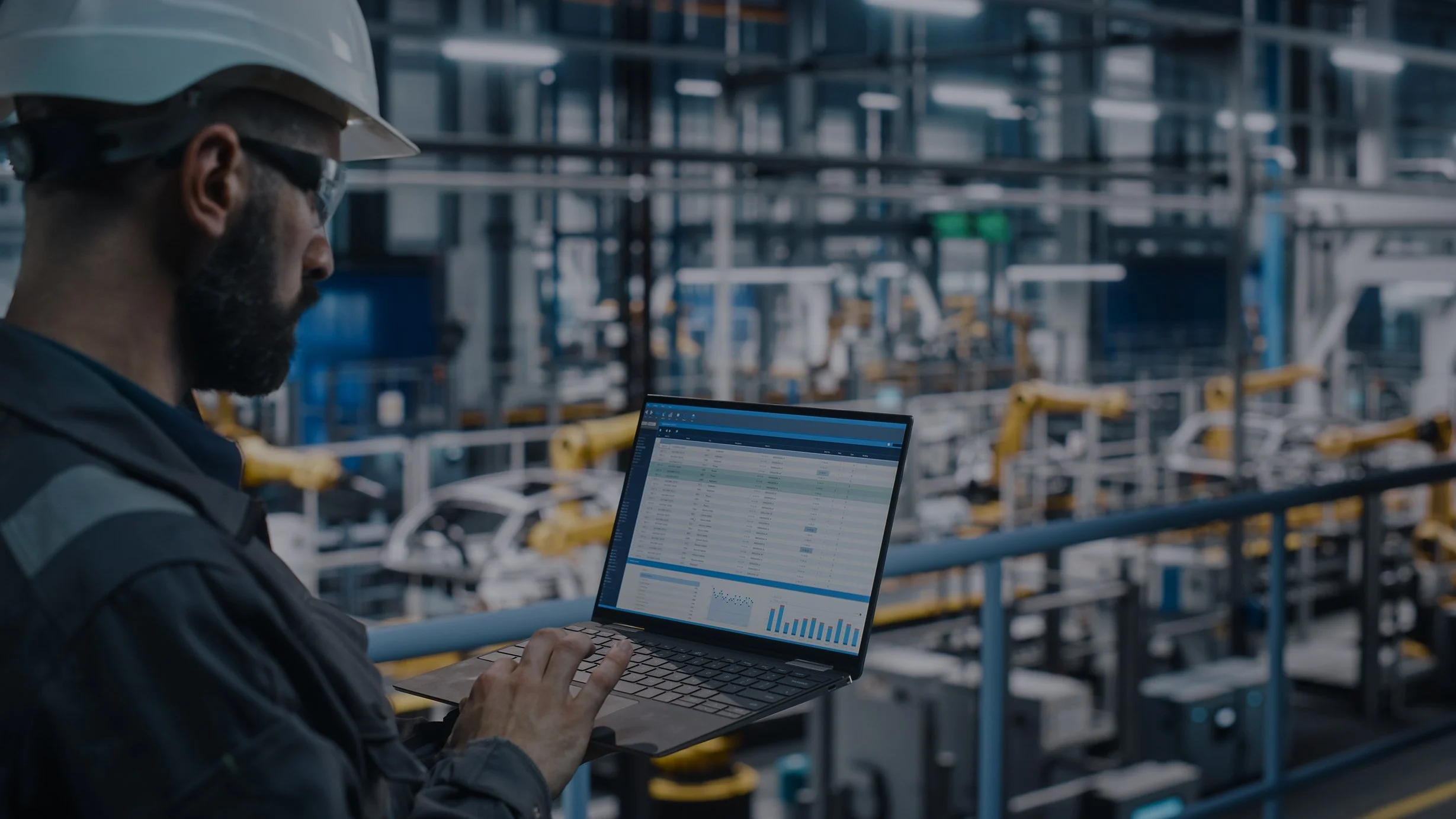 Industrial engineer in hard hat using a laptop to monitor automated factory machinery
