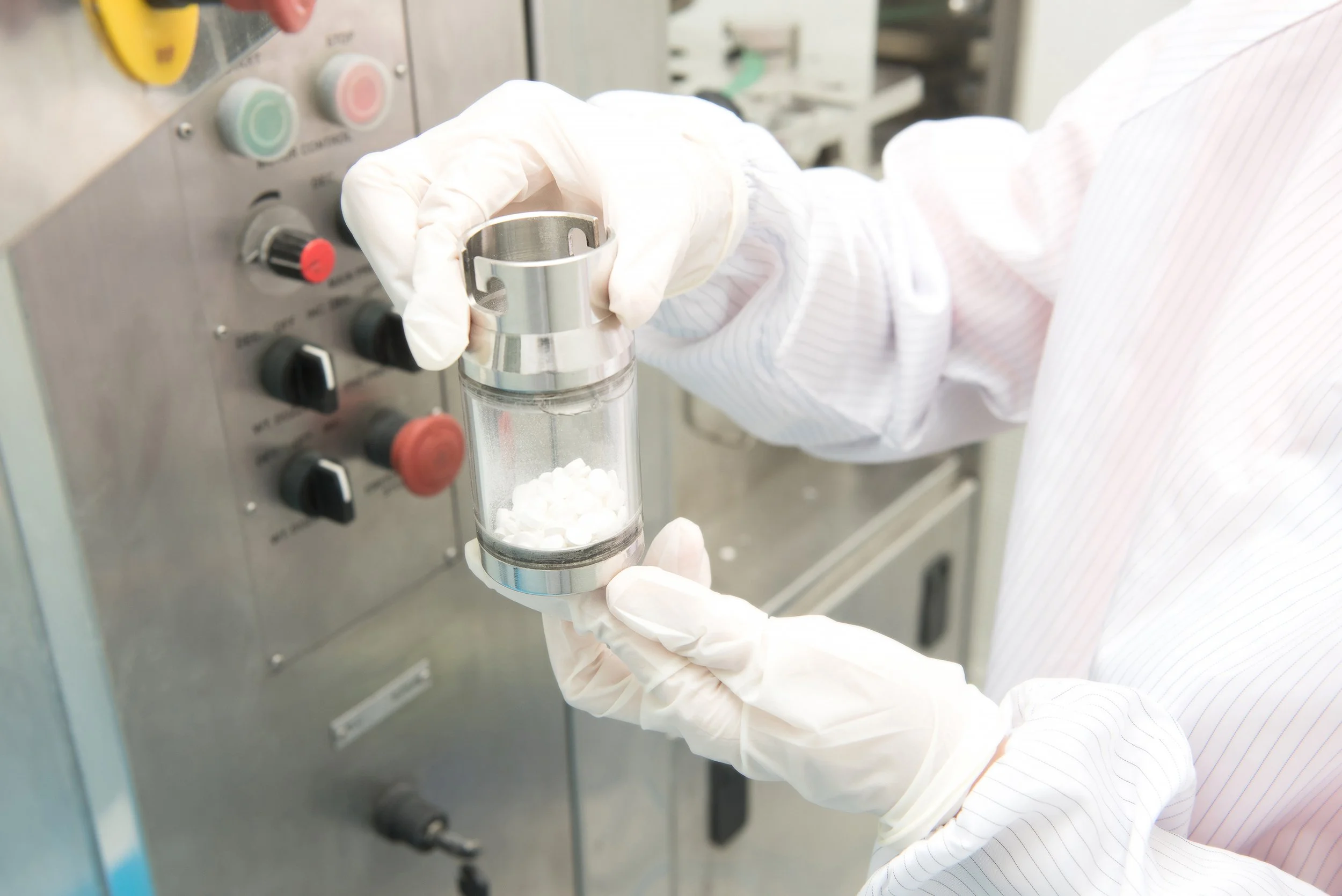 Gloved pharmaceutical worker inspecting white tablets in a stainless steel capsule-filling machine