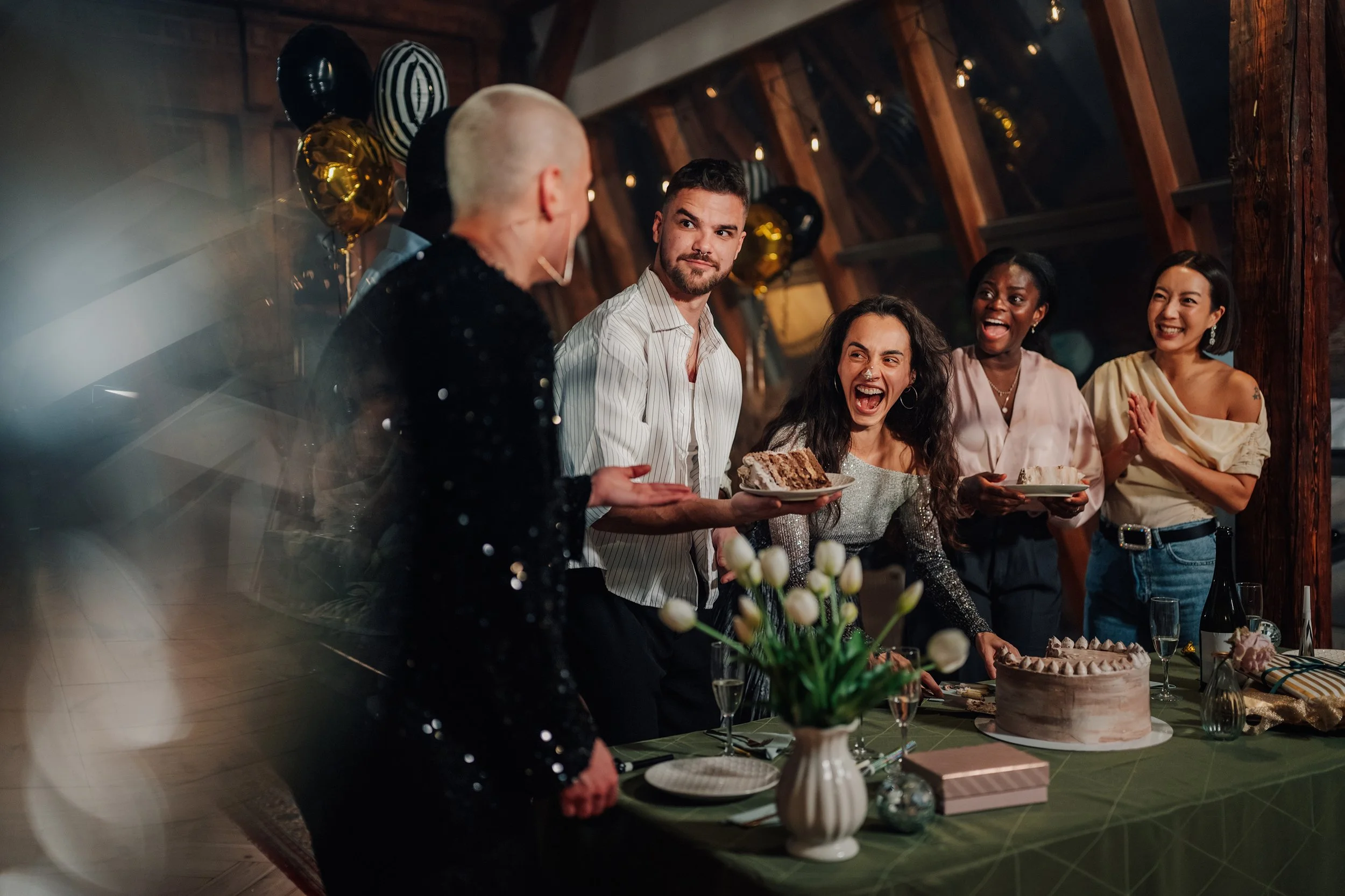 Friends celebrating around a dinner table with cake, champagne, balloons, and party decorations.