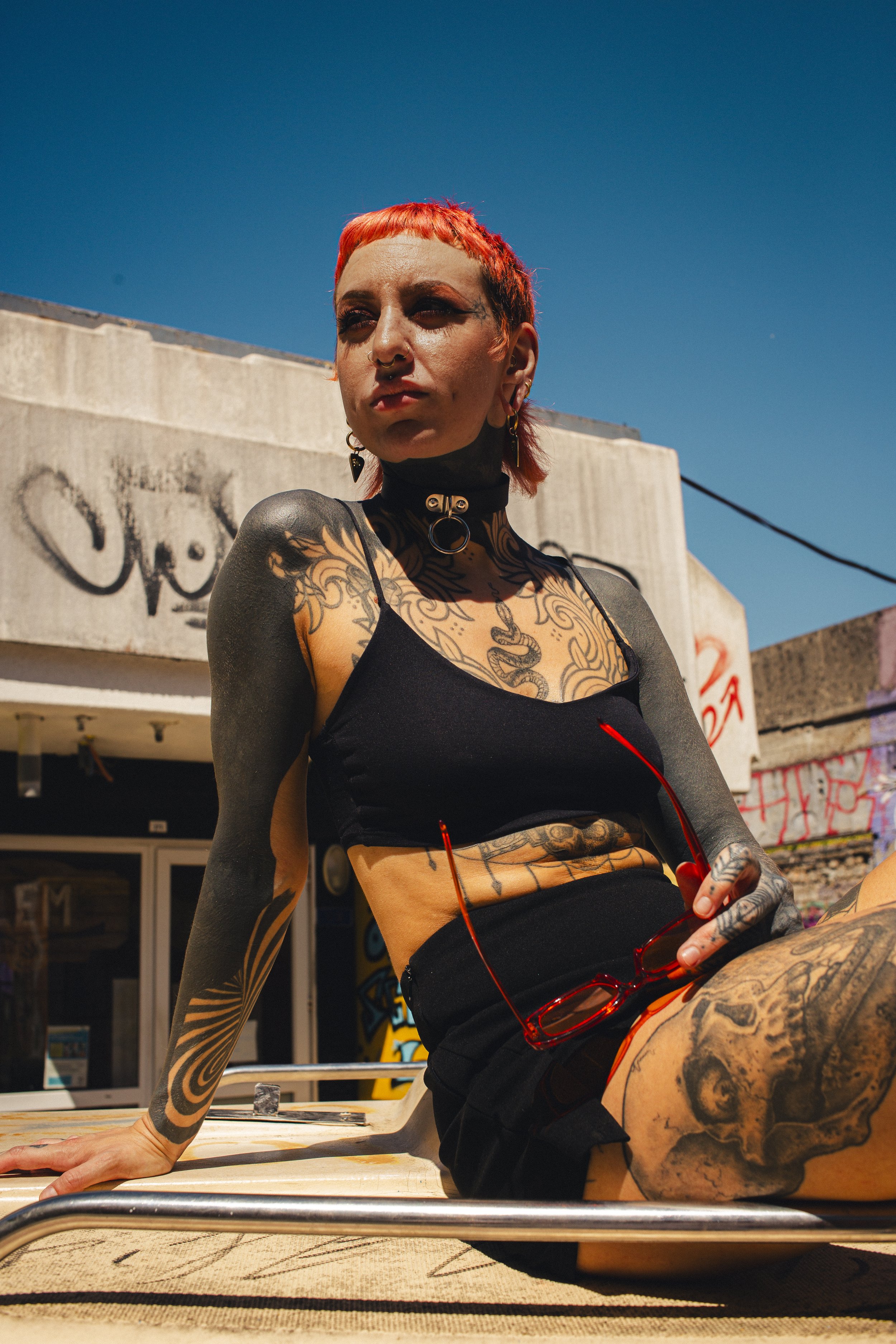Une femme tatouée avec des cheveux courts rouges foncés, portant un collier à œil de chat, un haut noir, et des lunettes rouges, posant à l'extérieur sous un ciel bleu clair.