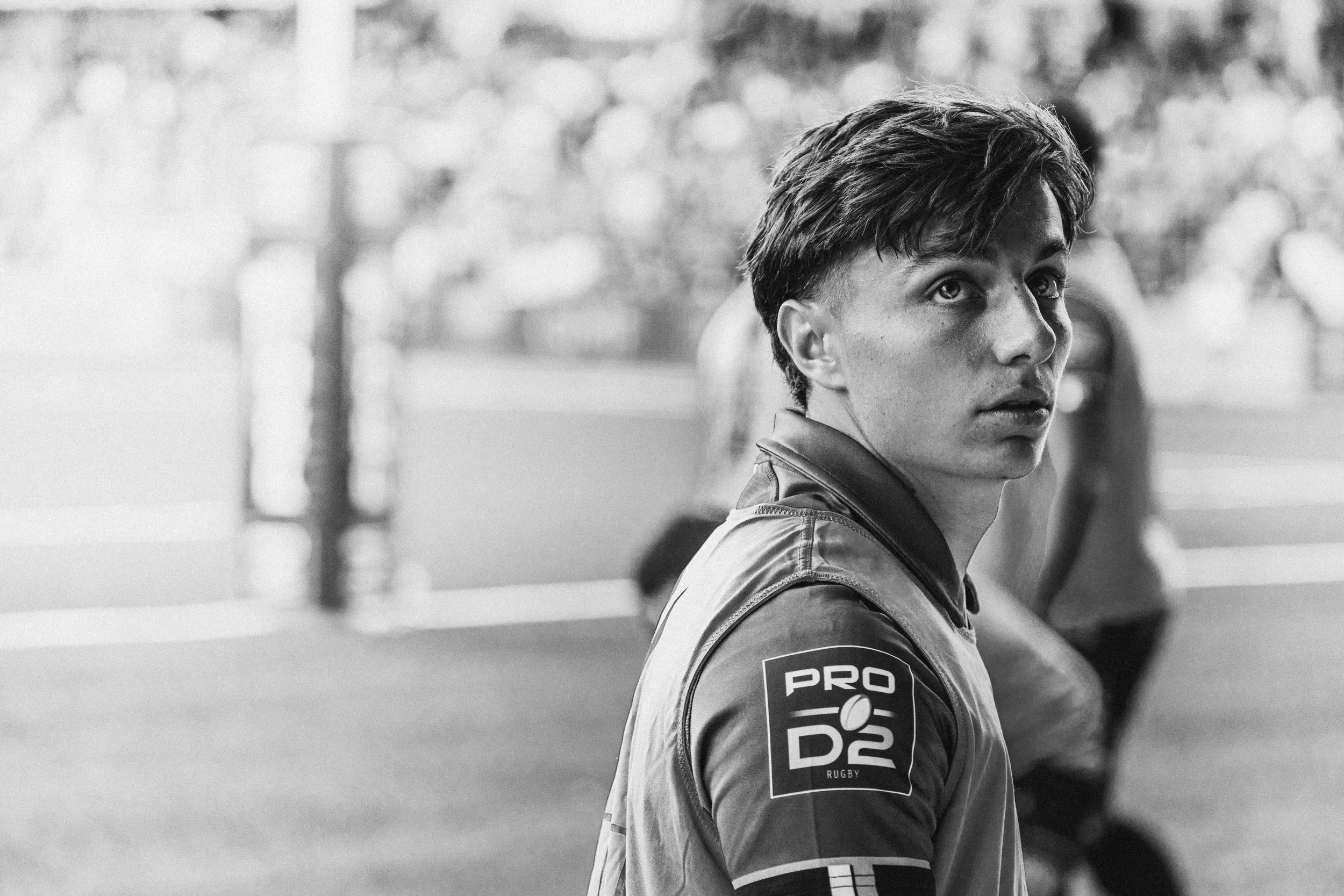 Un jeune homme portant un uniforme de rugby, regardant vers la droite, en noir et blanc, avec un arrière-plan flou.