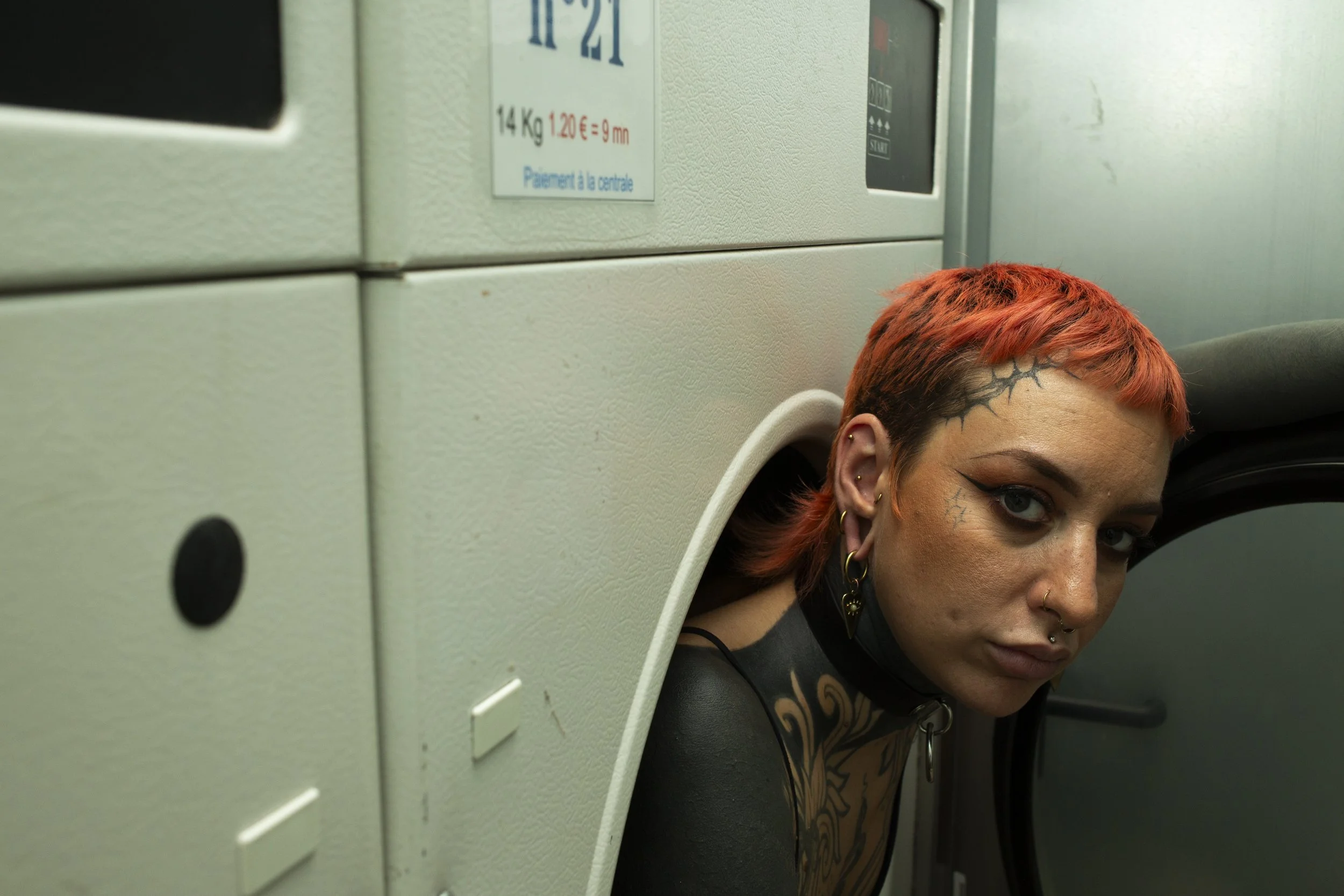 Une femme avec des cheveux orange et des tatouages sur le visage, regardant vers la caméra dans un endroit indéterminé, probablement près de machines ou de coffrets électriques.