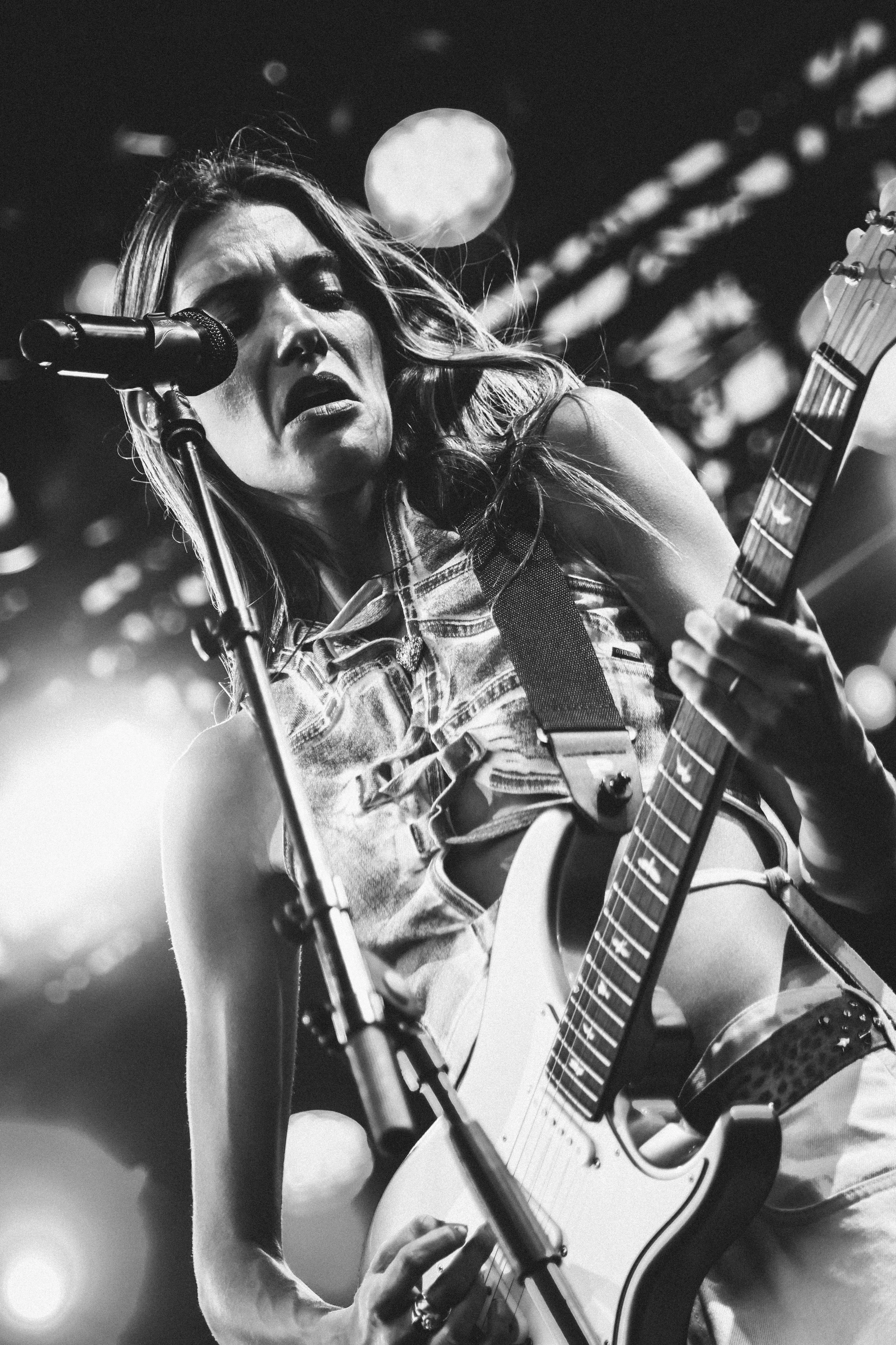 Une femme joue de la guitare électrique lors d'un concert, filmée en noir et blanc, avec un micro devant elle et un fond lumineux avec des formes floues.