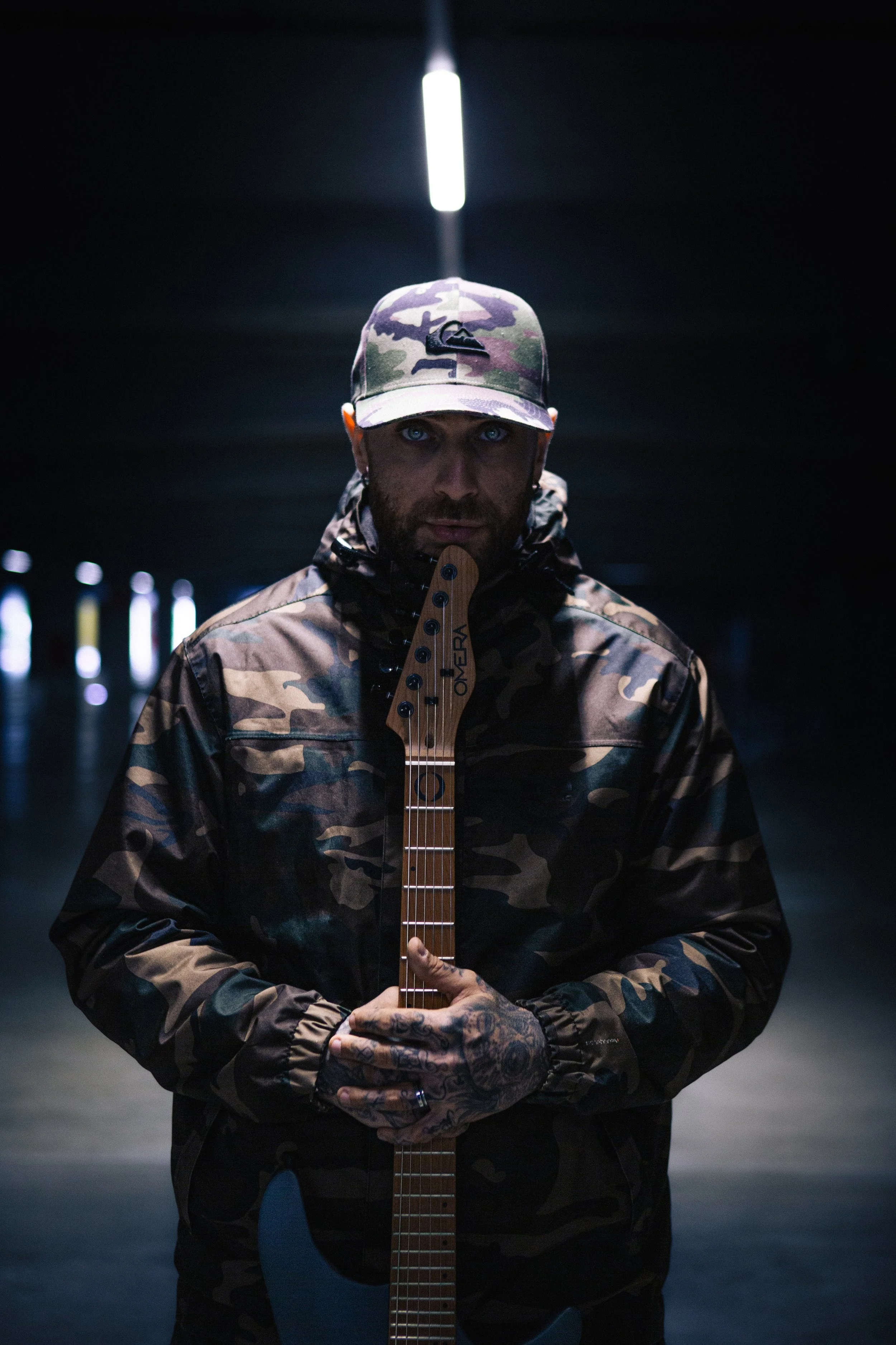Un homme portant un uniforme camouflage, un bonnet avec le logo Quiksilver, tenant une guitare acoustique devant un fond sombre et éclairé en haut.