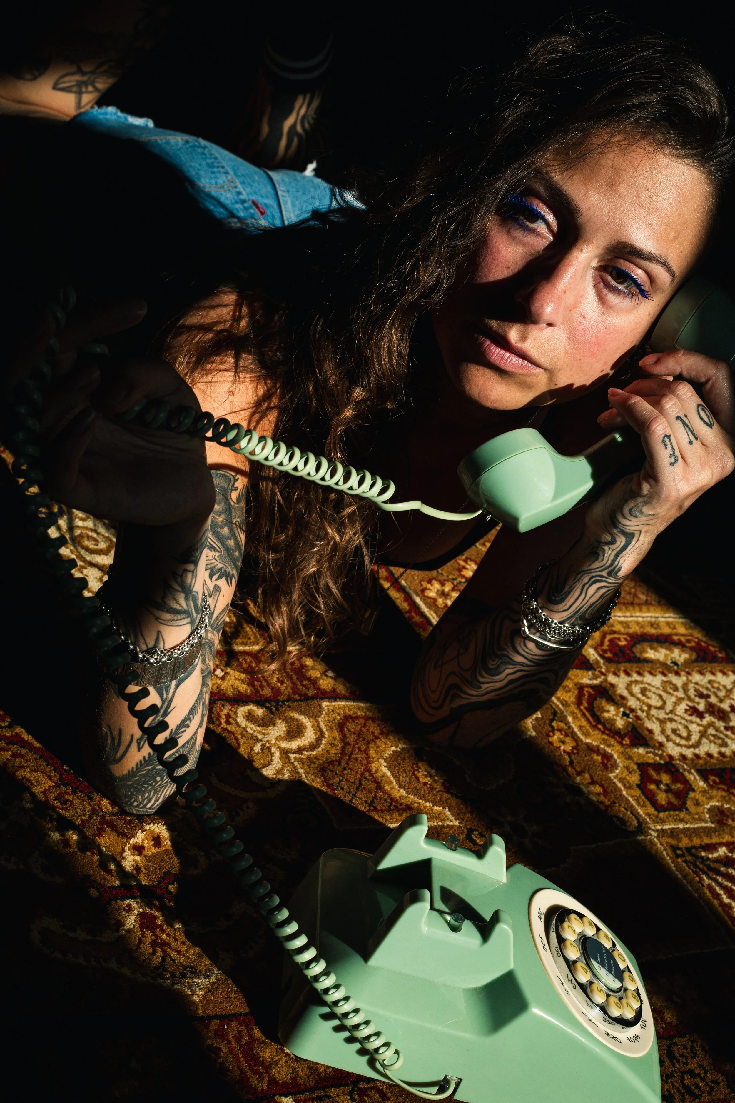 Jeune femme avec de nombreux tatouages, portant une veste en jean, à l'intérieur, en train de parler sur un téléphone Vintage de couleur vert pâle, assise sur un tapis à motif oriental.
