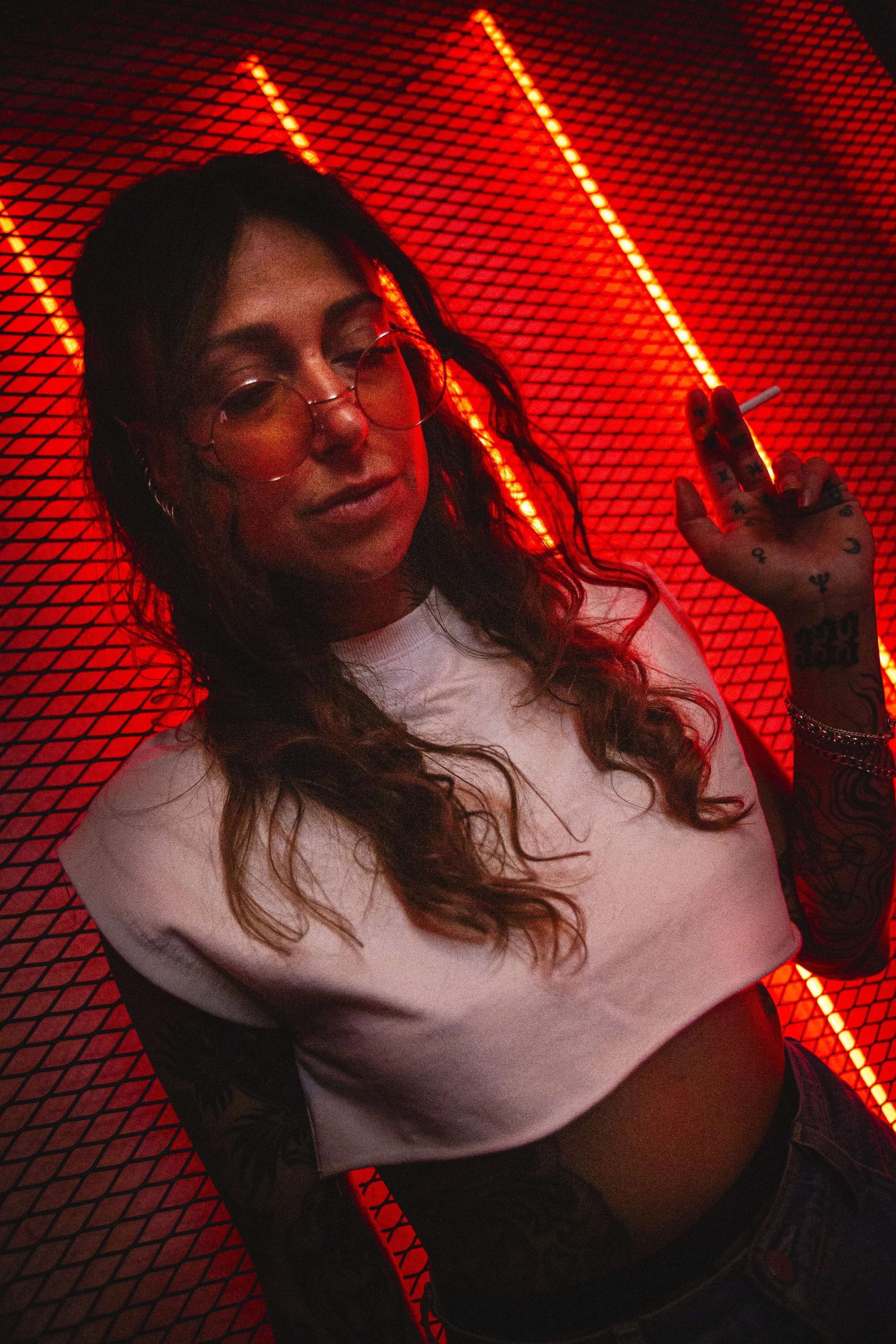 Femme avec lunettes rondes, pose dans un fond rouge lumineux, portant un t-shirt blanc et montrant une main tatouée avec une cigarette.
