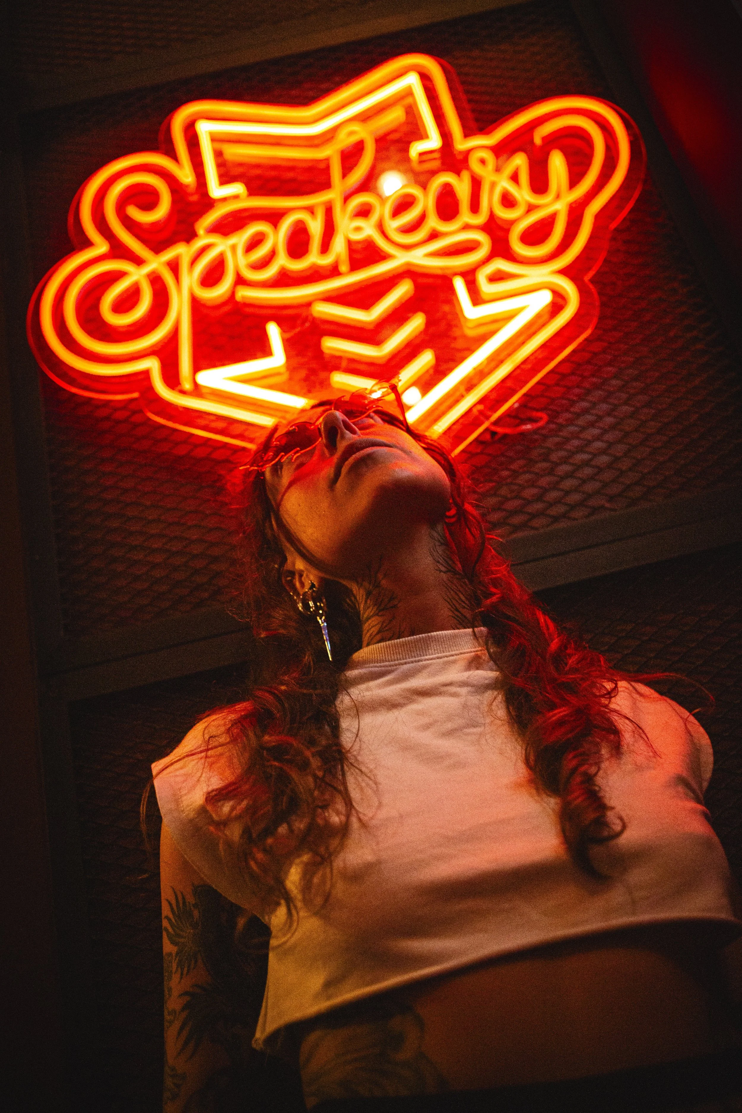 Une personne avec de longs cheveux roux, portant des lunettes de soleil, un t-shirt blanc et un tatouage sur le bras, se trouve sous un panneau lumineux rouge avec le mot "Speakeasy" en néon.