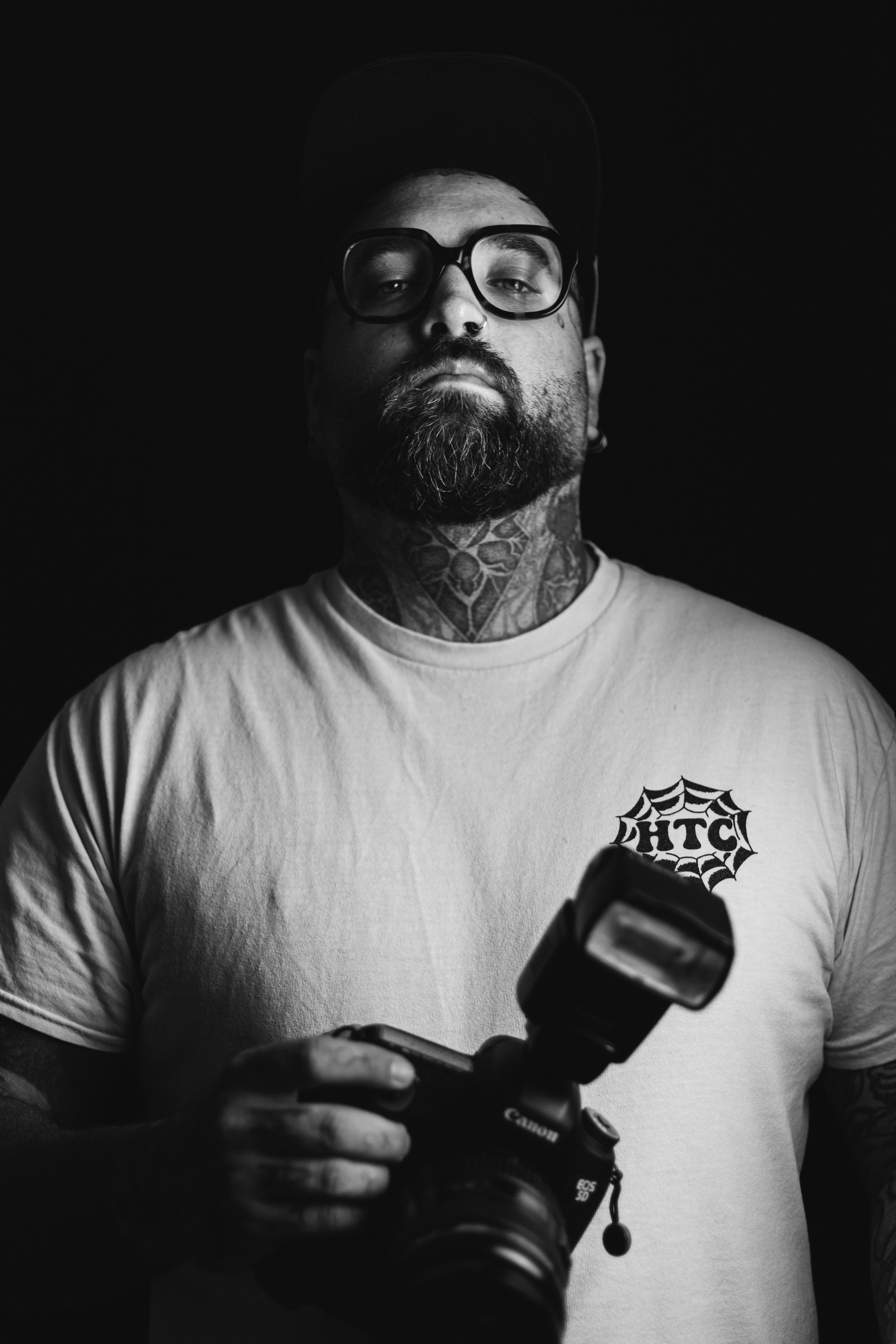 Un homme portant des lunettes et un t-shirt blanc avec un logo de cuisine, tenant un appareil photo numérique, regardant vers l'objectif, avec un fond noir.