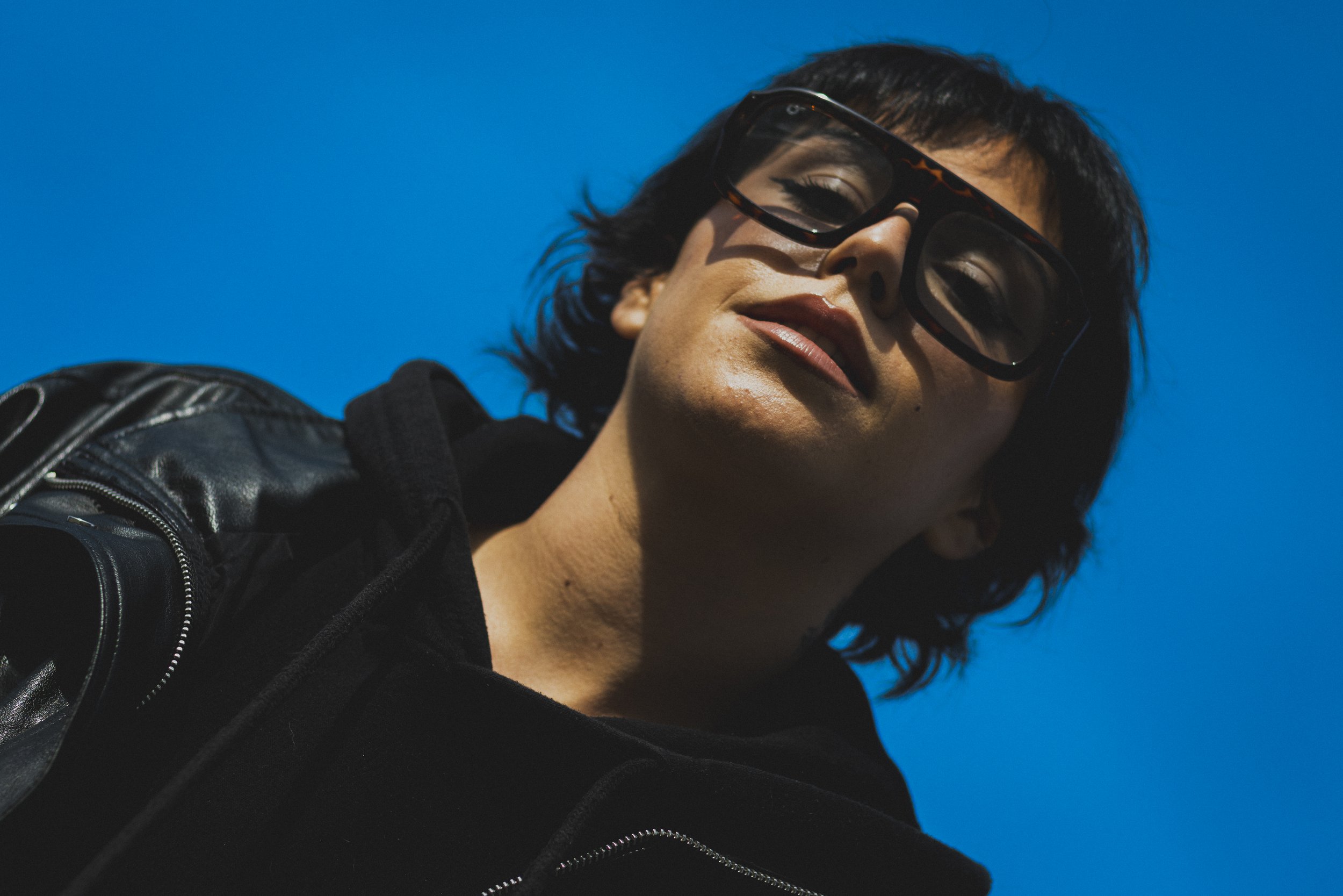 Une femme avec des cheveux courts noirs, portant des lunettes à tortoise, souriant légèrement, portant une veste en cuir noir et un sweat noir, avec un fond bleu clair.