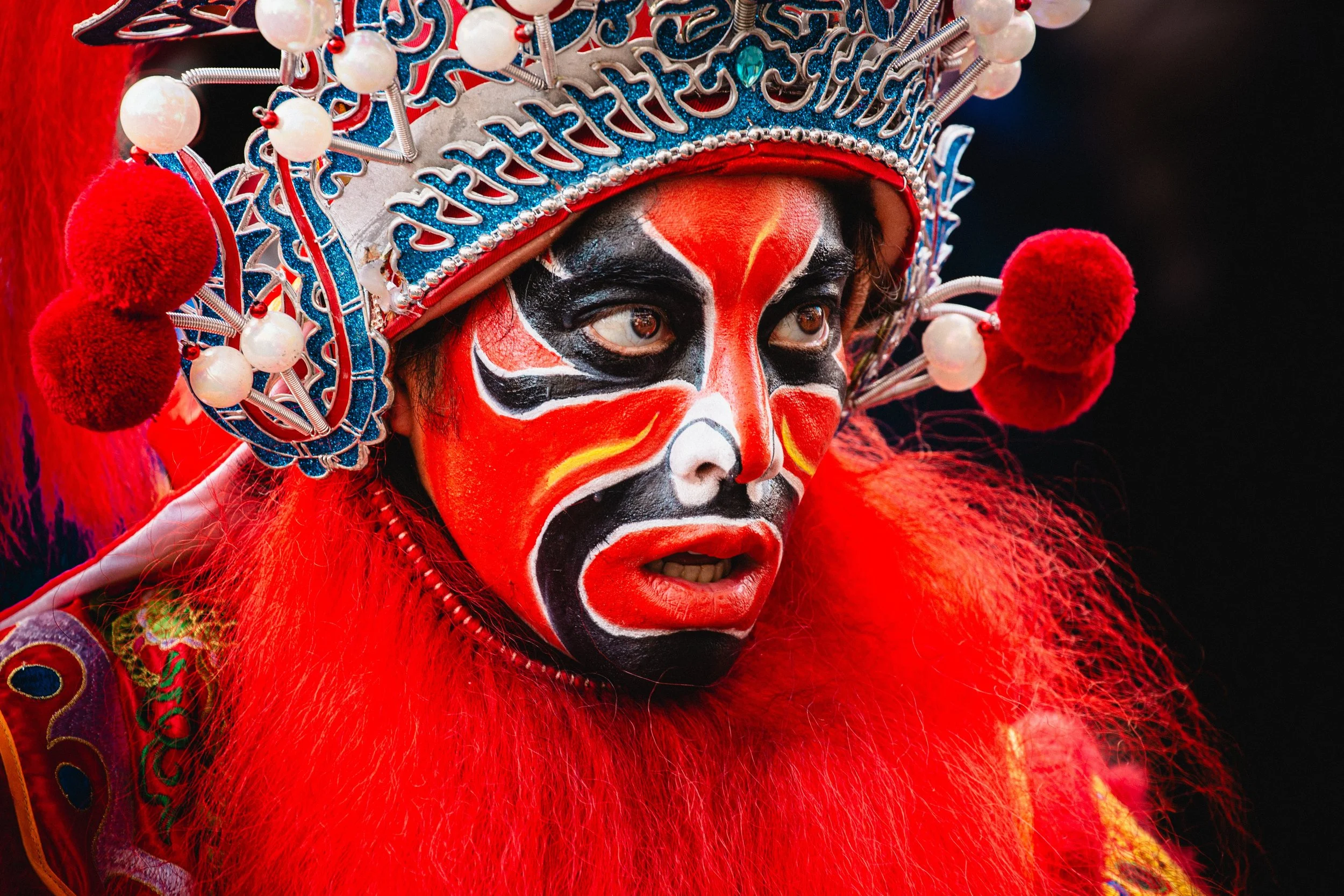 Personne déguisée en personnage traditionnel, avec un maquillage facial coloré en rouge, noir, blanc et jaune, portant un costume extravagant avec un couvre-chef orné de perles, de pompons rouges, et de détails en bleu et argent.