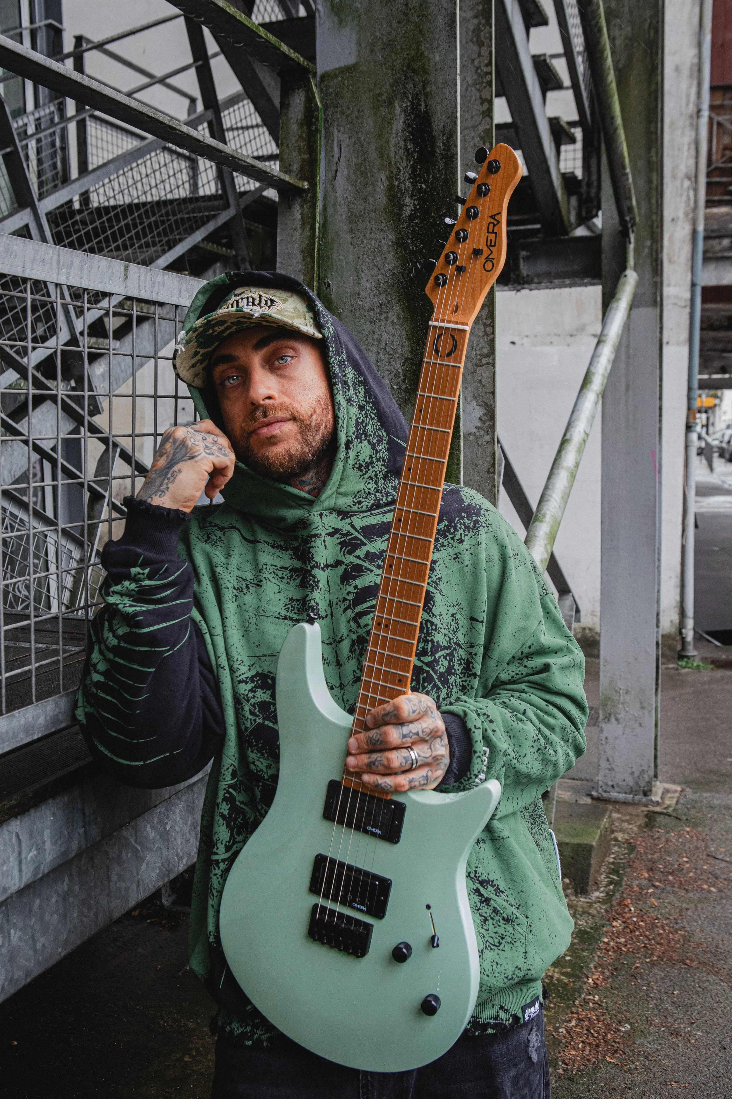 Un homme avec des tatouages, portant une capuche verte et un chapeau camouflage, pose avec une guitare électrique blanche dans un environnement urbain industriel.